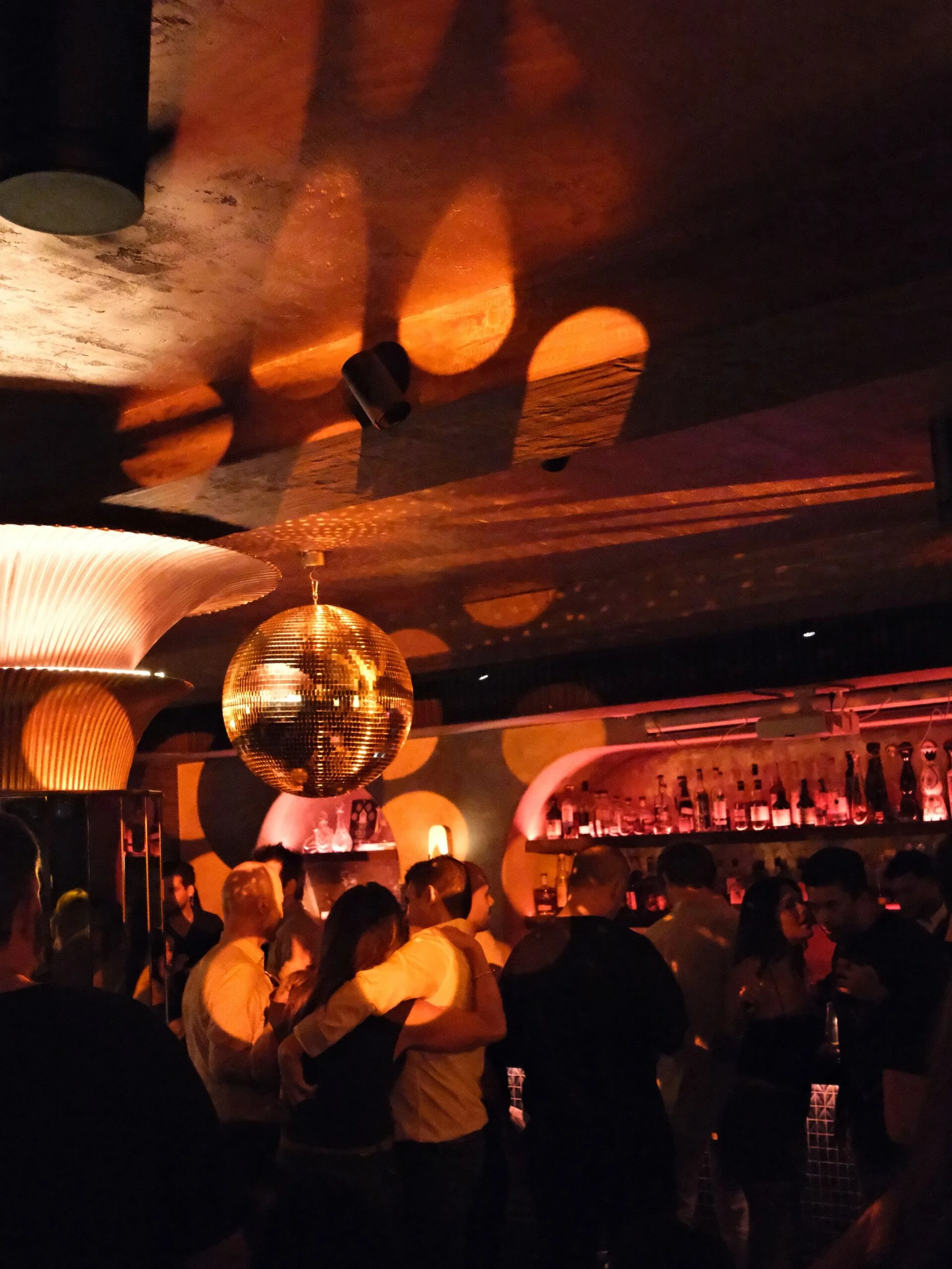 Dimly lit bar or nightclub with a crowd of people, gold disco ball, and unique ceiling lighting creating circular glow effects.