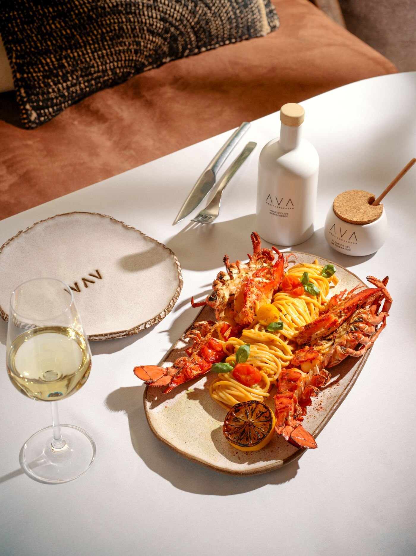 A scenic plate of lobster and spaghetti with cherry tomatoes and basil on a ceramic platter, a glass of white wine, and a white table setting with cutlery and a white bottle of olive oil, on a white table with a brown cushioned bench in the background.