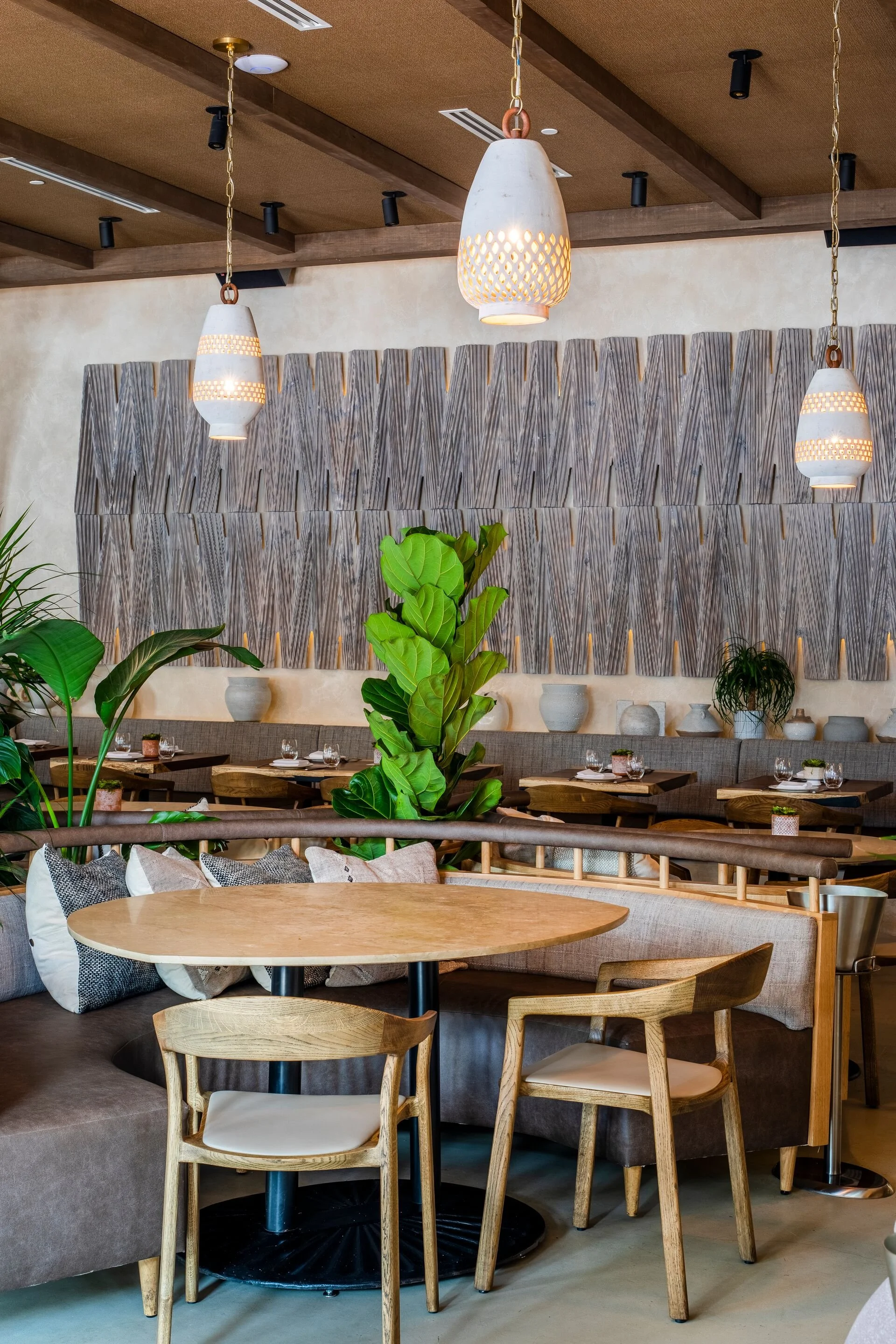 An indoor dining area with wooden tables and chairs, decorative plants, pendant lights, and a textured wall art piece.