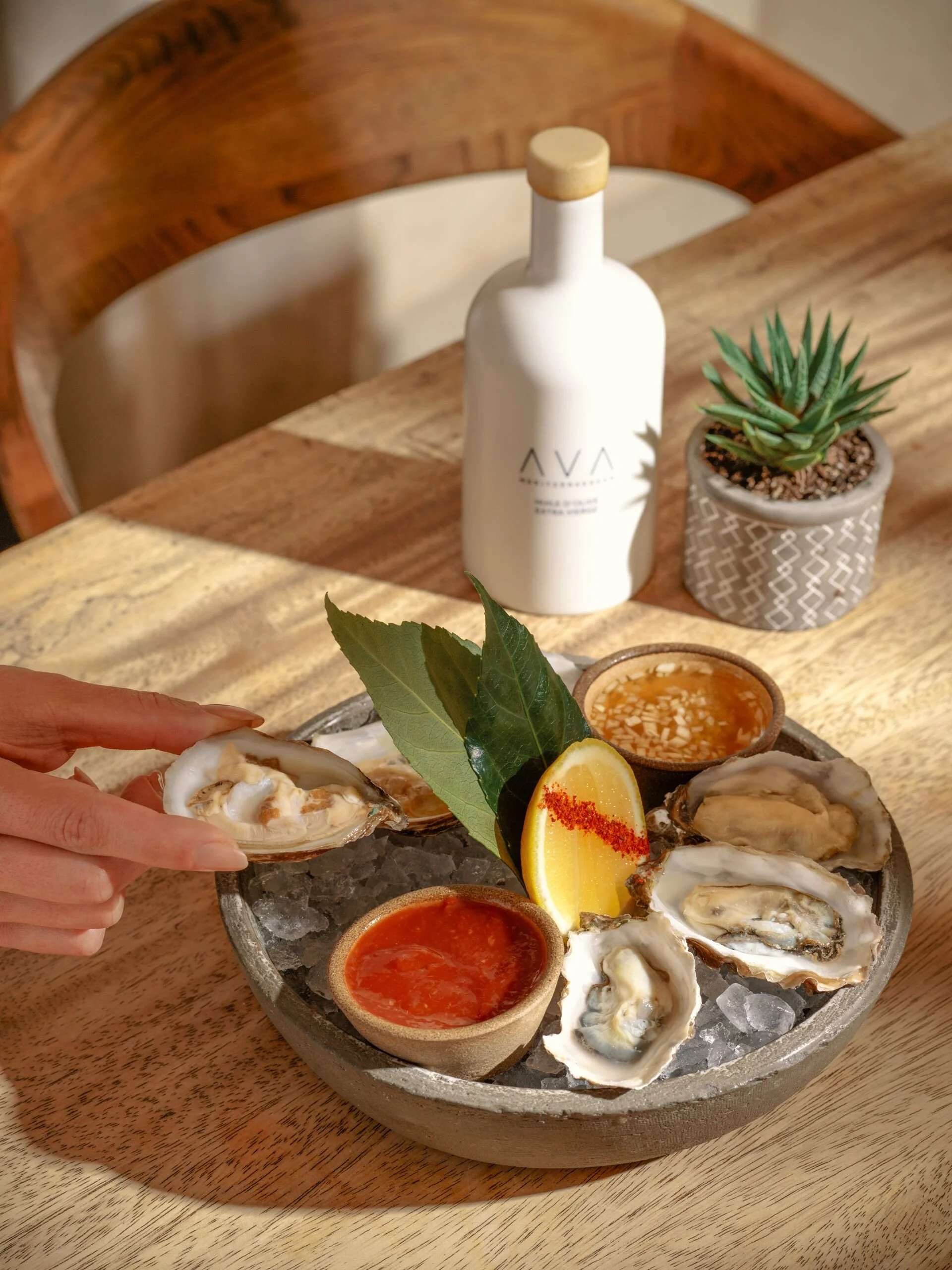 A person is holding an oyster shell with oysters on a bed of ice, alongside a lemon wedge with red seasoning, and two small bowls of dipping sauces on a wooden table.