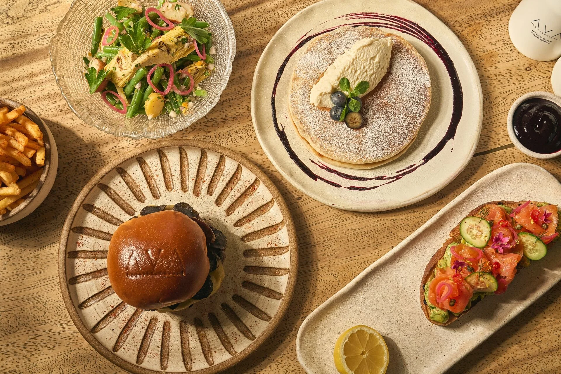 A variety of dishes including a salad with greens and pink onions, a stack of pancakes with whipped cream and blueberries, a burger, and a veggie toast with cucumber and carrot slices on a wooden table.