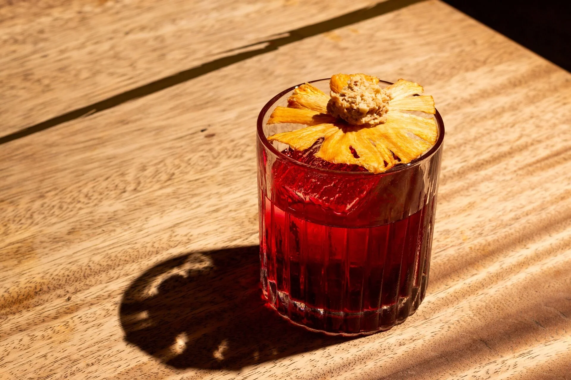 A tall glass of bright red beverage garnished with a dried pineapple slice and a scoop of what appears to be white frothy topping, placed on a wooden surface with a shadow cast across the tabletop.