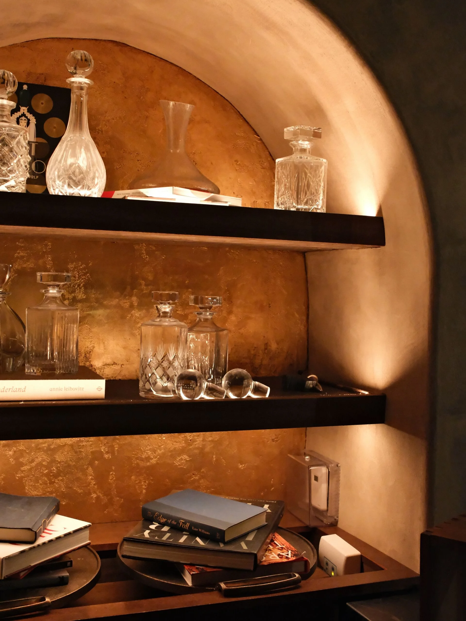 A set of shelves with various glass decanters and liquor bottles, some books, and a black table with more books and objects in front of a textured brown wall.