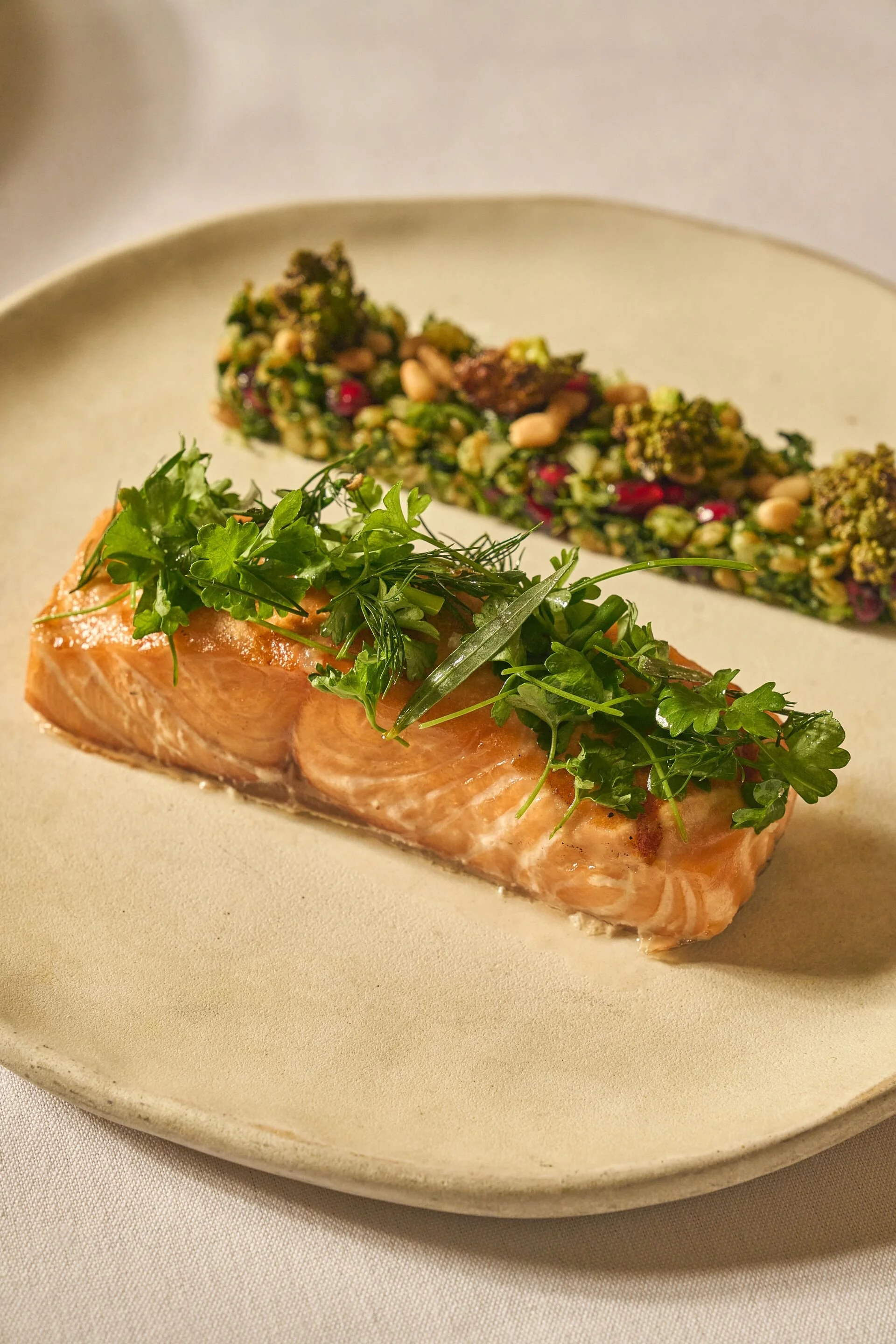 A plate with a piece of cooked salmon garnished with fresh parsley and herbs, and a side of broccoli and vegetable salad.