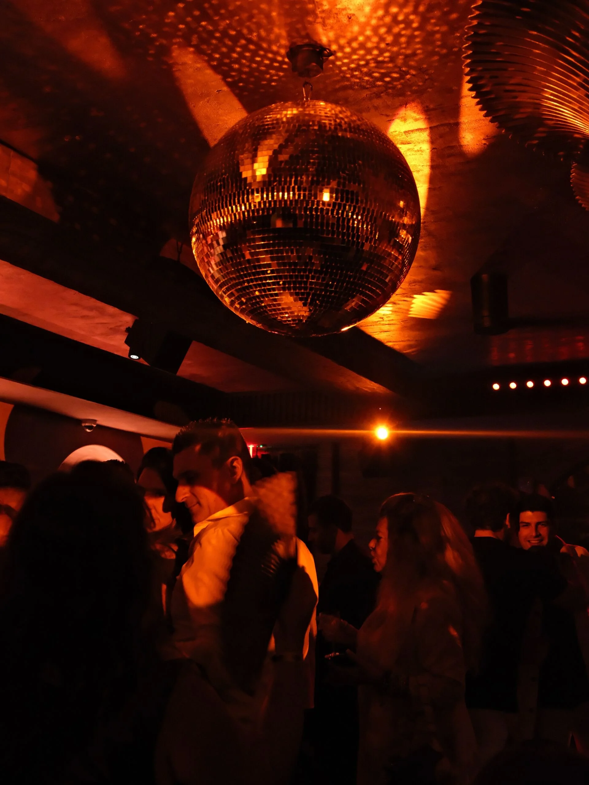 People dancing and socializing in a dimly lit nightclub with a large disco ball hanging from the ceiling, reflecting orange light.