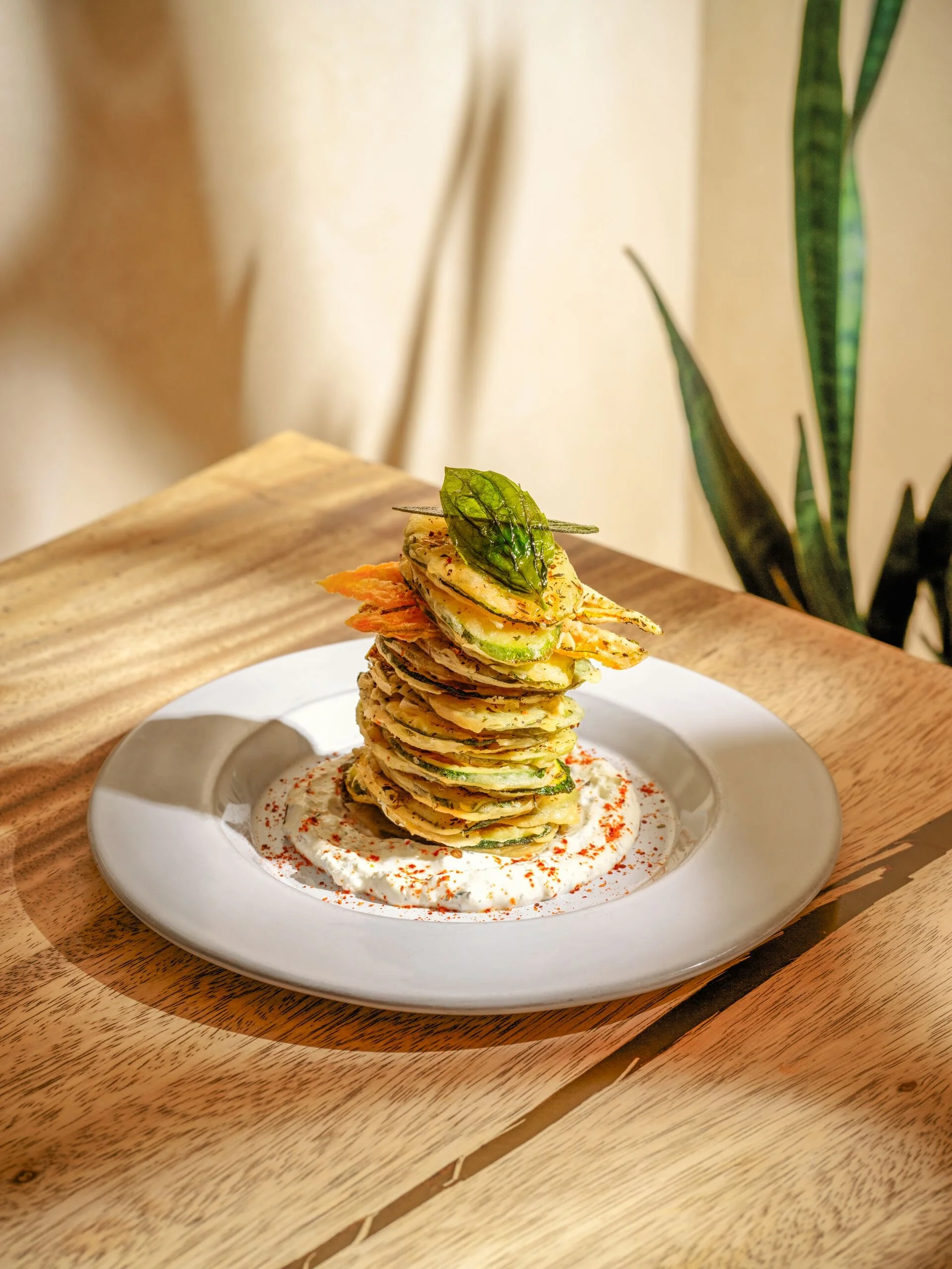A plated dish with multiple layered pieces of grilled vegetables topped with a green leaf, served on a white plate with a bed of sauce and sprinkled seasoning, placed on a wooden tray.