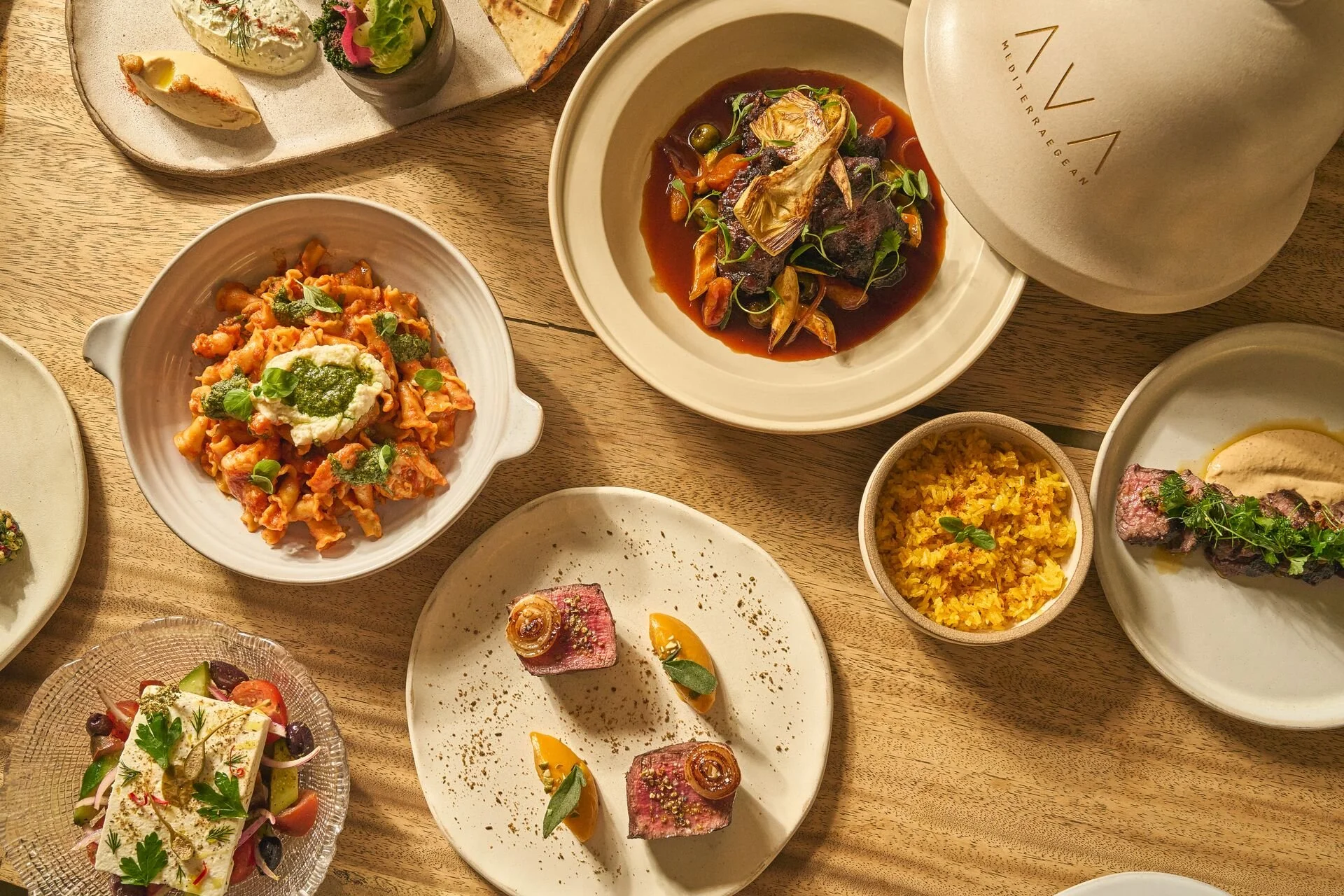 A variety of plated dishes and small bowls of food on a wooden table, including pasta, salad, rice, roasted vegetables, and meat.