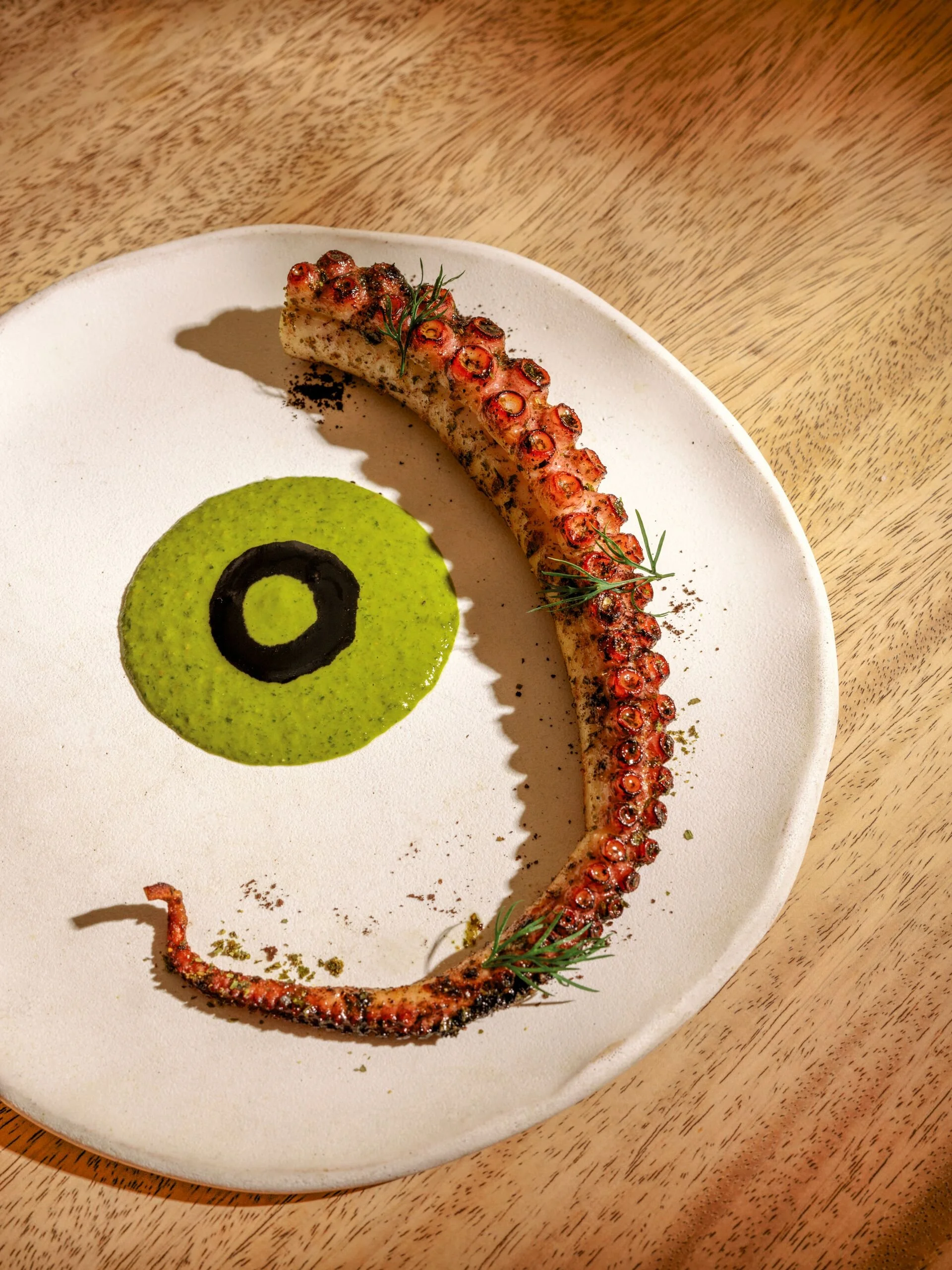 Grilled octopus tentacle garnished with herbs, served on a white plate with a green sauce and black decorative circle, on a wooden table.
