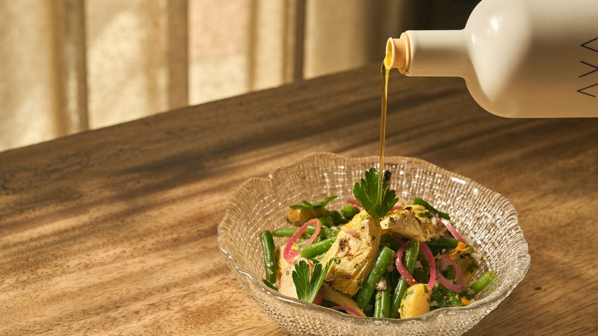A glass bowl of green bean salad topped with fresh parsley, chicken, and pink pickled onions, with dressing being poured over it from a white bottle.