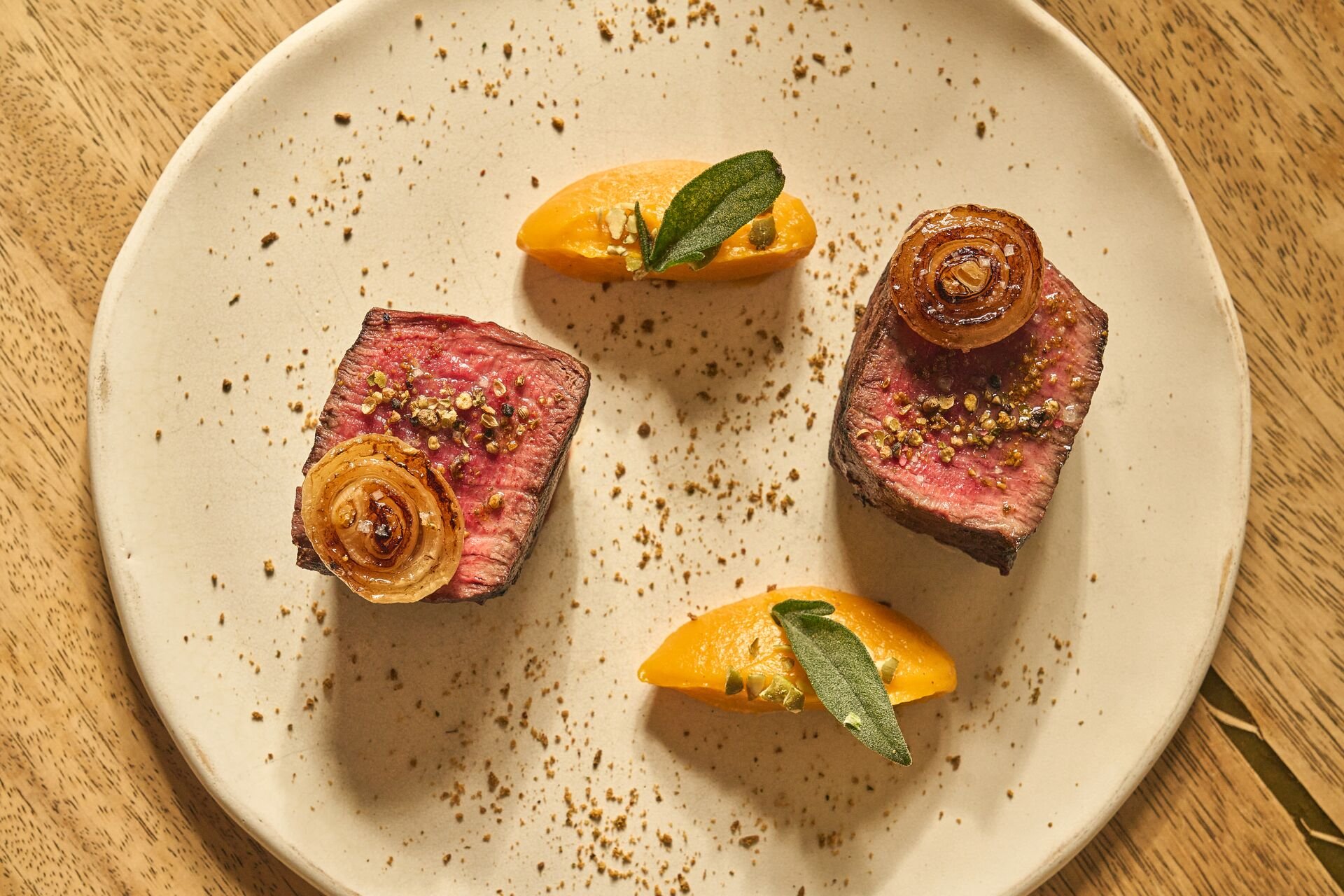Two pieces of medium-rare beef tenderloin topped with caramelized onions and sprinkled with black pepper, garnished with a small green leaf, on a white plate with a yellow sauce and crumbs, served on a wooden table.