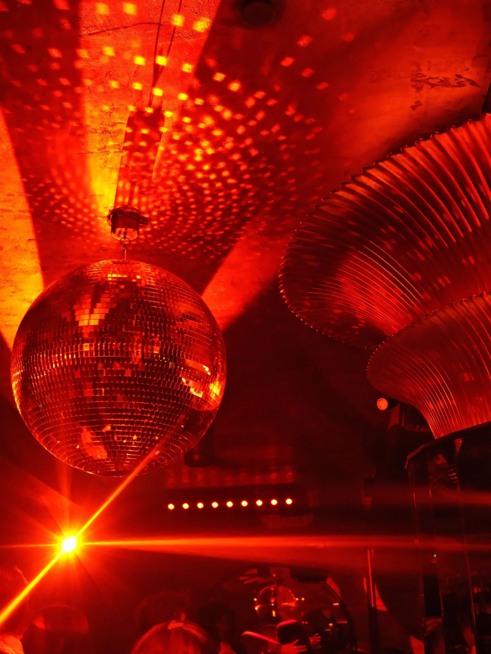 A dark room illuminated with red light, featuring a disco ball casting dotted patterns on the ceiling and walls, with a bright yellow light source in the bottom left corner. People are visible at the bottom of the image, enjoying the atmosphere.