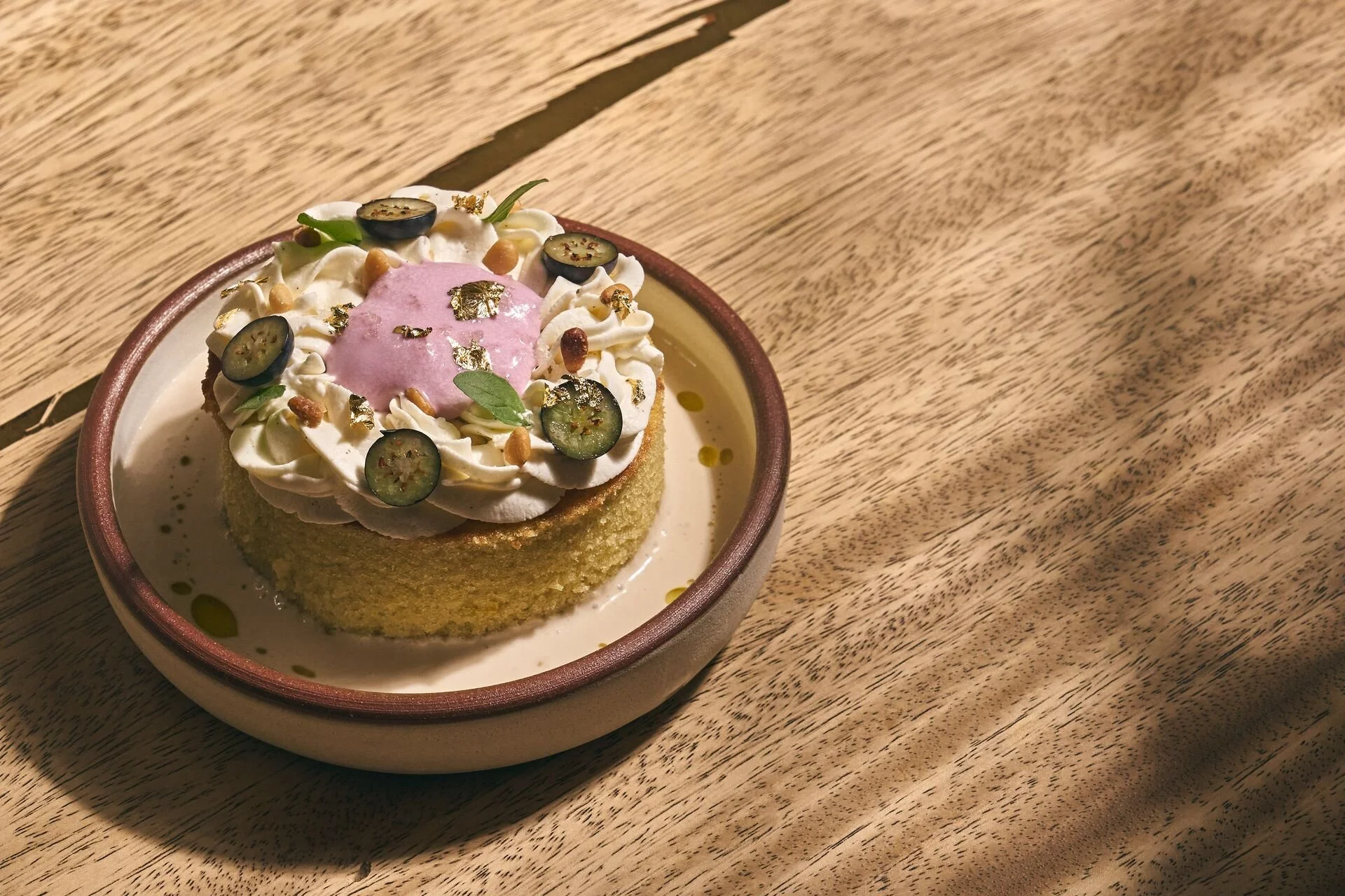 A dessert in a small bowl, topped with whipped cream, purple sauce, sliced blueberries, small green leaves, gold leaf flakes, and small golden and brown toppings, on a wooden surface.
