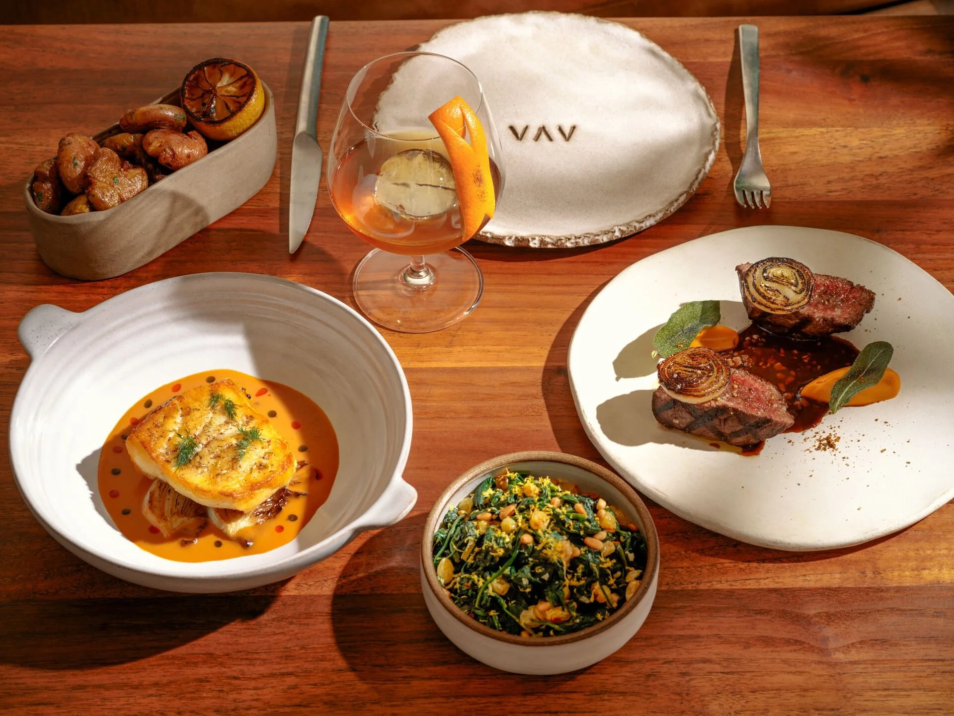 A variety of dishes including a grilled meat entrée with vegetables, a baked fish fillet in orange sauce, a bowl of greens with corn and herbs, a bowl of seasoned potatoes, and a cocktail with lemon and orange garnishes on a wooden table.