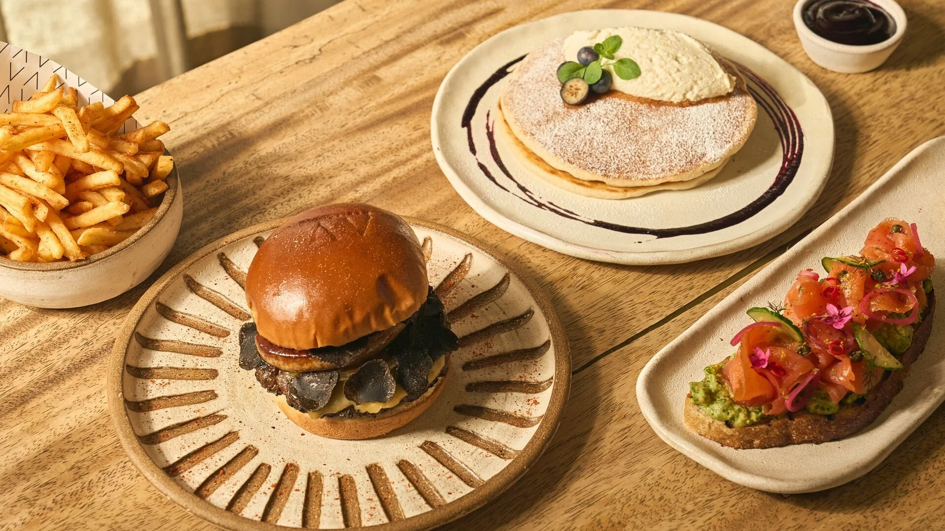 Assorted dishes on a wooden table including French fries, a cheeseburger with truffle chips, a plate of pancakes with berries and whipped cream, and an avocado toast with smoked salmon and edible flowers.