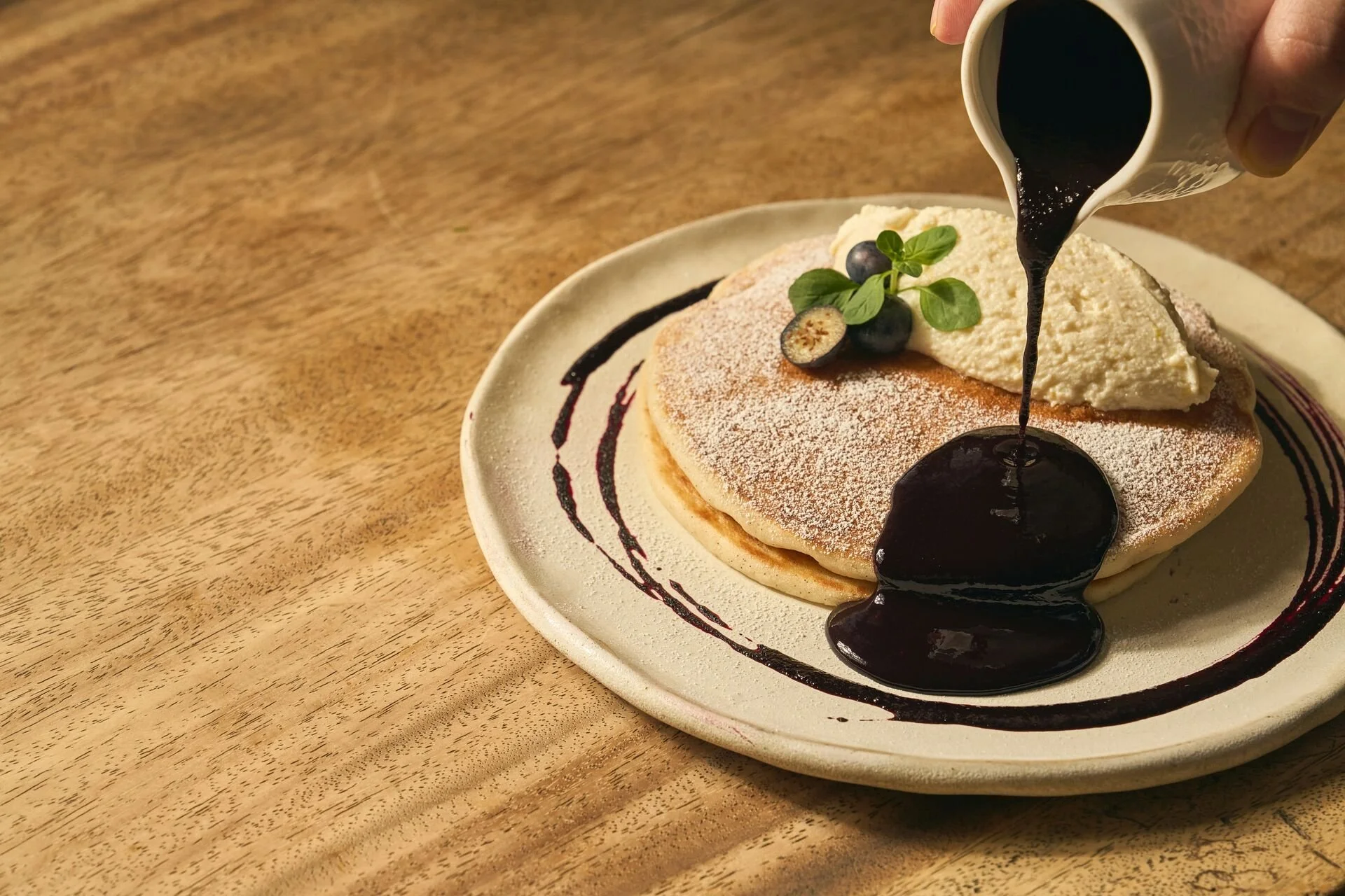 Plate with stacks of pancakes dusted with powdered sugar, topped with a dollop of whipped cream, blueberries, and a sprig of mint, with syrup being poured over them.