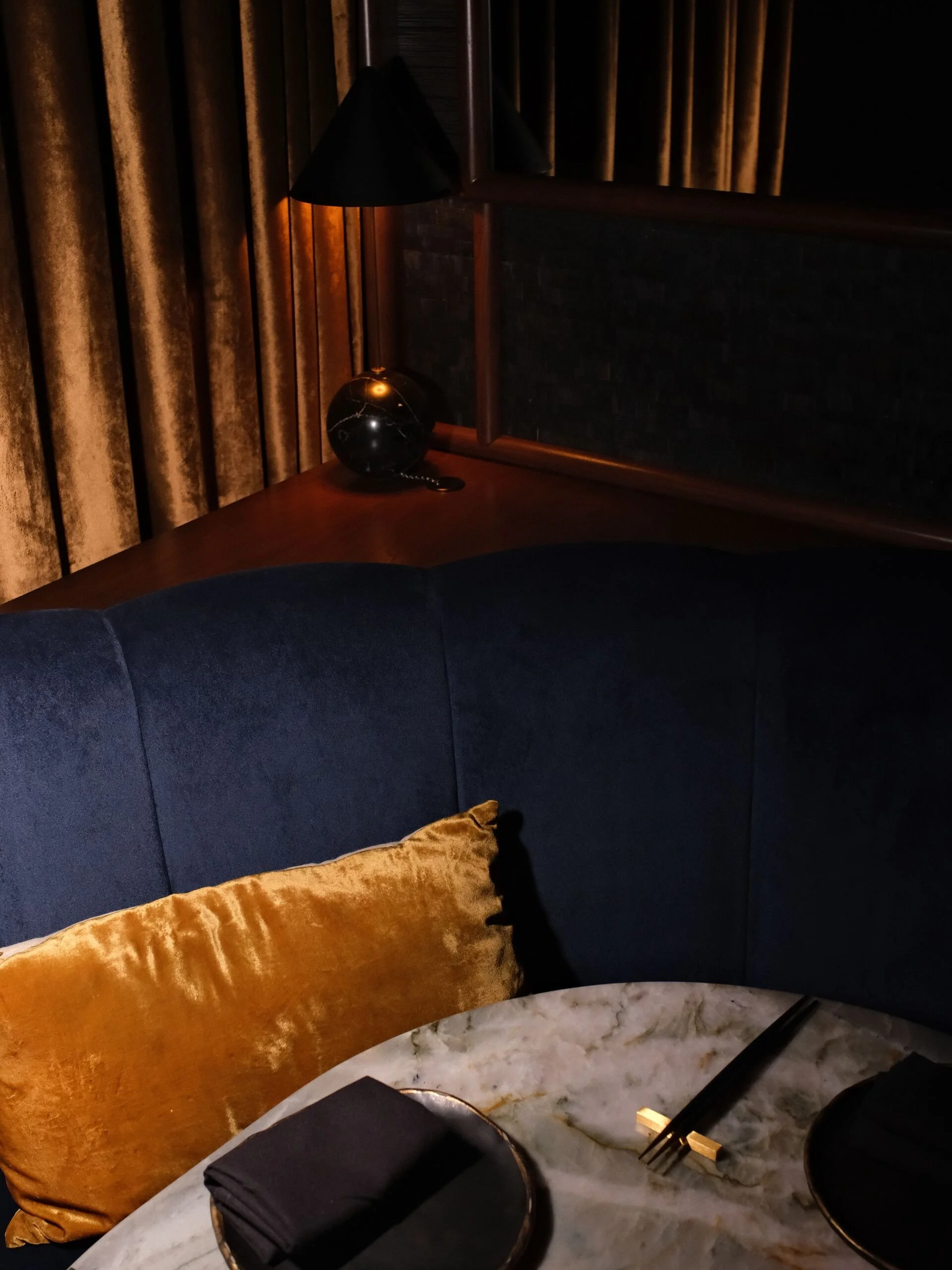A dark bedroom corner with a navy blue velvet sofa, a golden cushion, a marble table, a black handbag, a black fork, and a gold-handled black plate on the table.