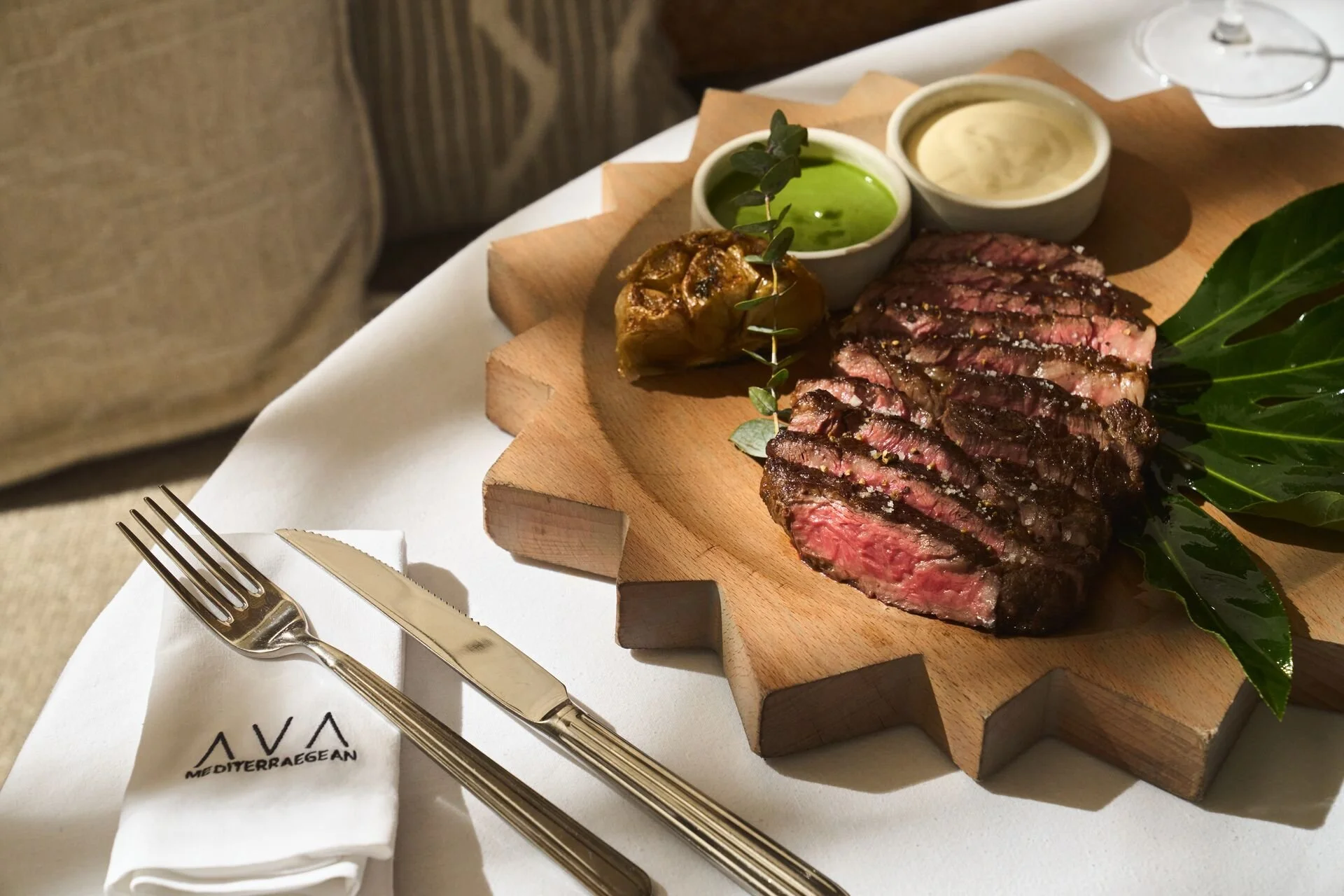 Slices of cooked steak served on a wooden cutting board with green leaves, accompanied by two small bowls of sauce and a roasted garlic clove. A fork and knife are placed on a white napkin with the restaurant's logo.