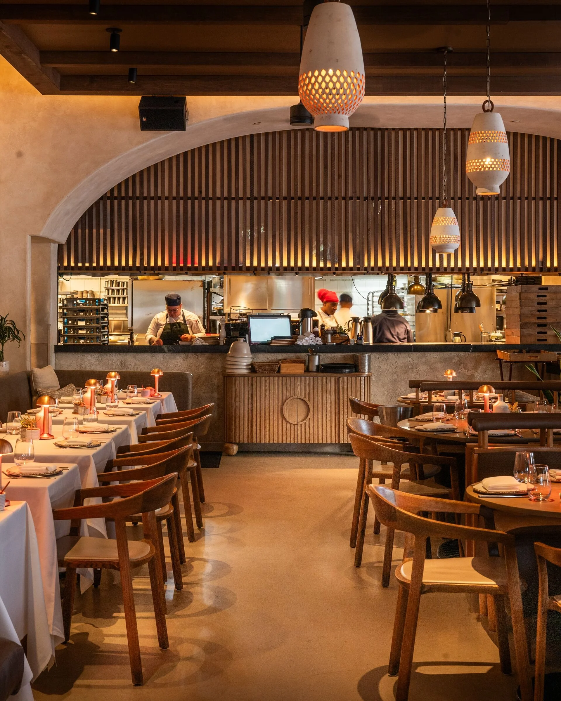 Interior of a restaurant with dining tables set with white tablecloths, glasses, plates, and cutlery, and wooden chairs. The background features a kitchen area with chefs working, framed by a wooden arched ceiling and hanging pendant lights.