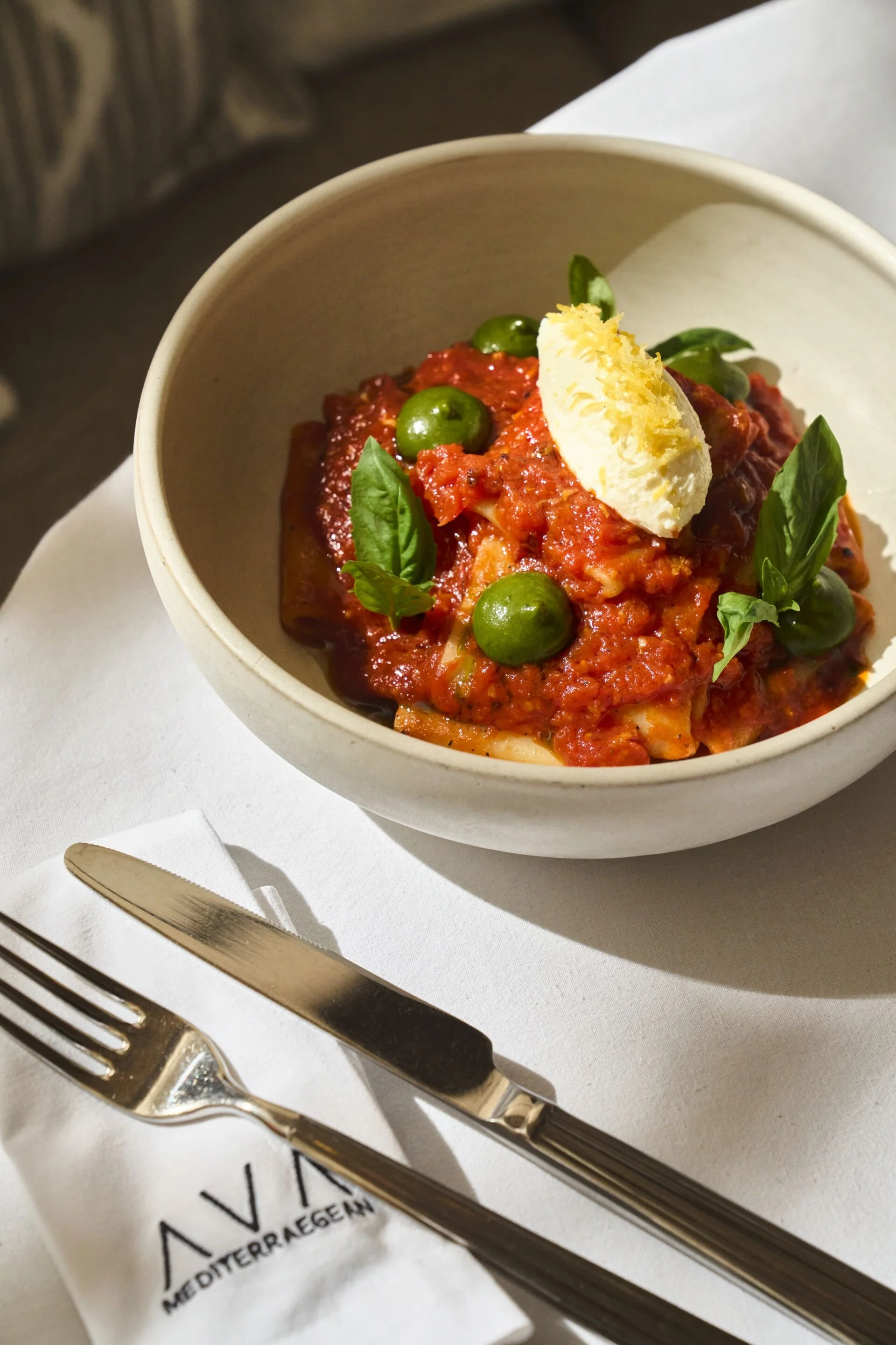 A bowl of pasta with tomato sauce, green peas, basil leaves, and a dollop of ricotta cheese on top, placed on a white table with a fork and knife, and a napkin that has text partially visible.