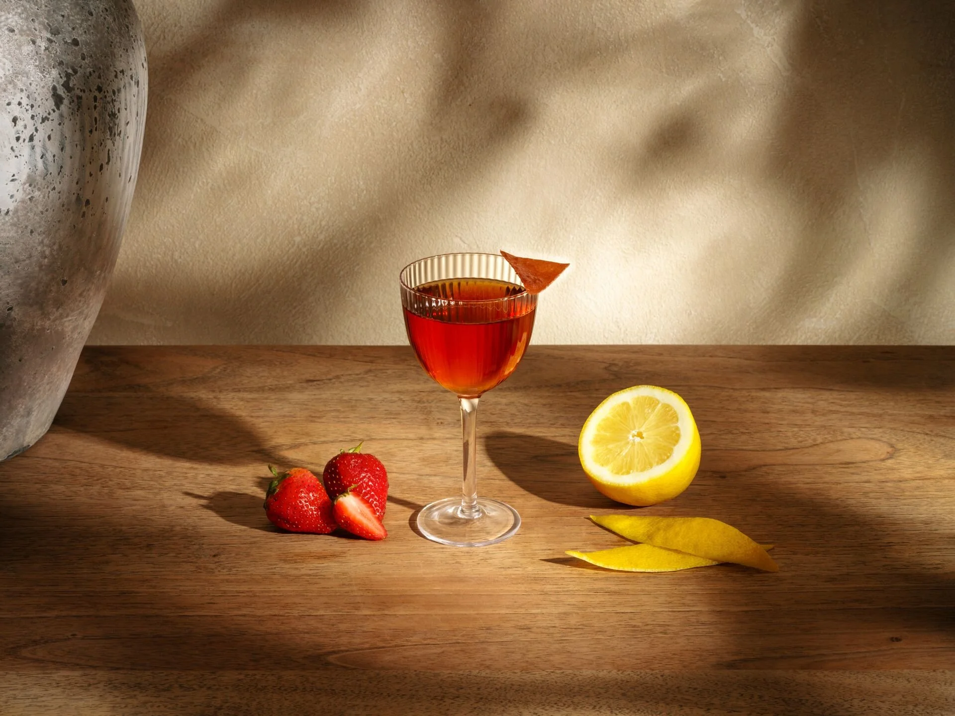 A glass of red cocktail with a lemon twist garnish, surrounded by strawberries, lemon half, and lemon peel slices on a wooden surface, with a textured background.
