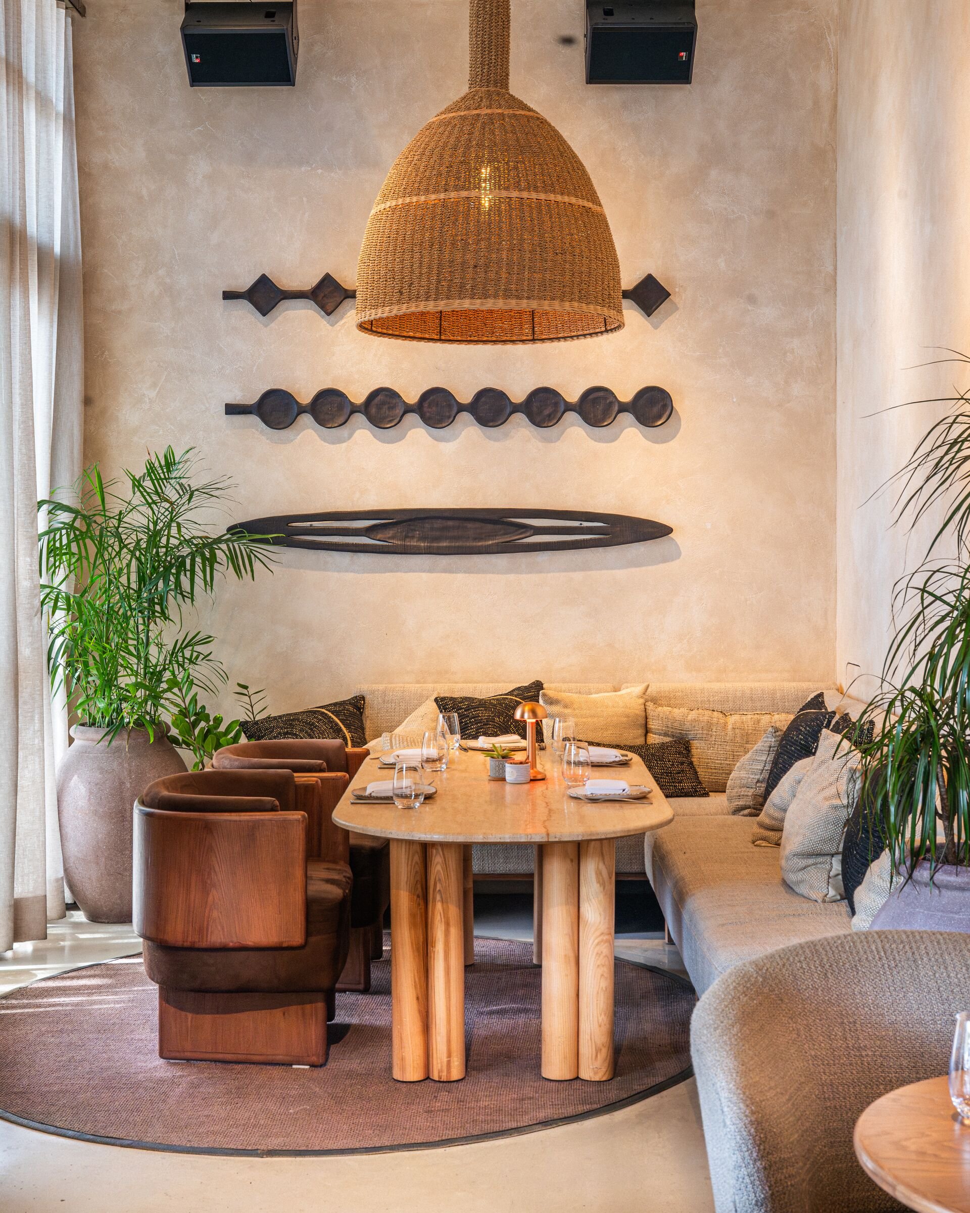 A cozy restaurant interior with a wooden table, surrounded by chairs and a beige sofa with patterned pillows. A large woven pendant light hangs above the table, and potted plants add greenery. The wall features decorative black wall art and two mount