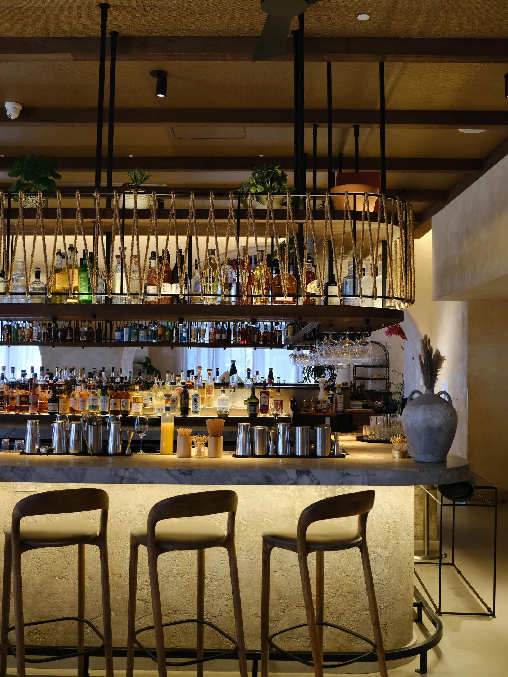 A bar area with a marble countertop, wooden chairs, and a two-tier shelf holding liquor bottles. The background includes decorative plants and a subdued lighting ambiance.