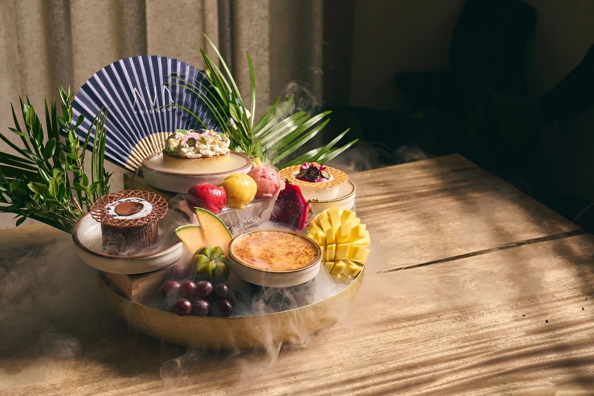 An assortment of desserts on a round tray, decorated with green leaves and a blue fan, placed on a wooden table.