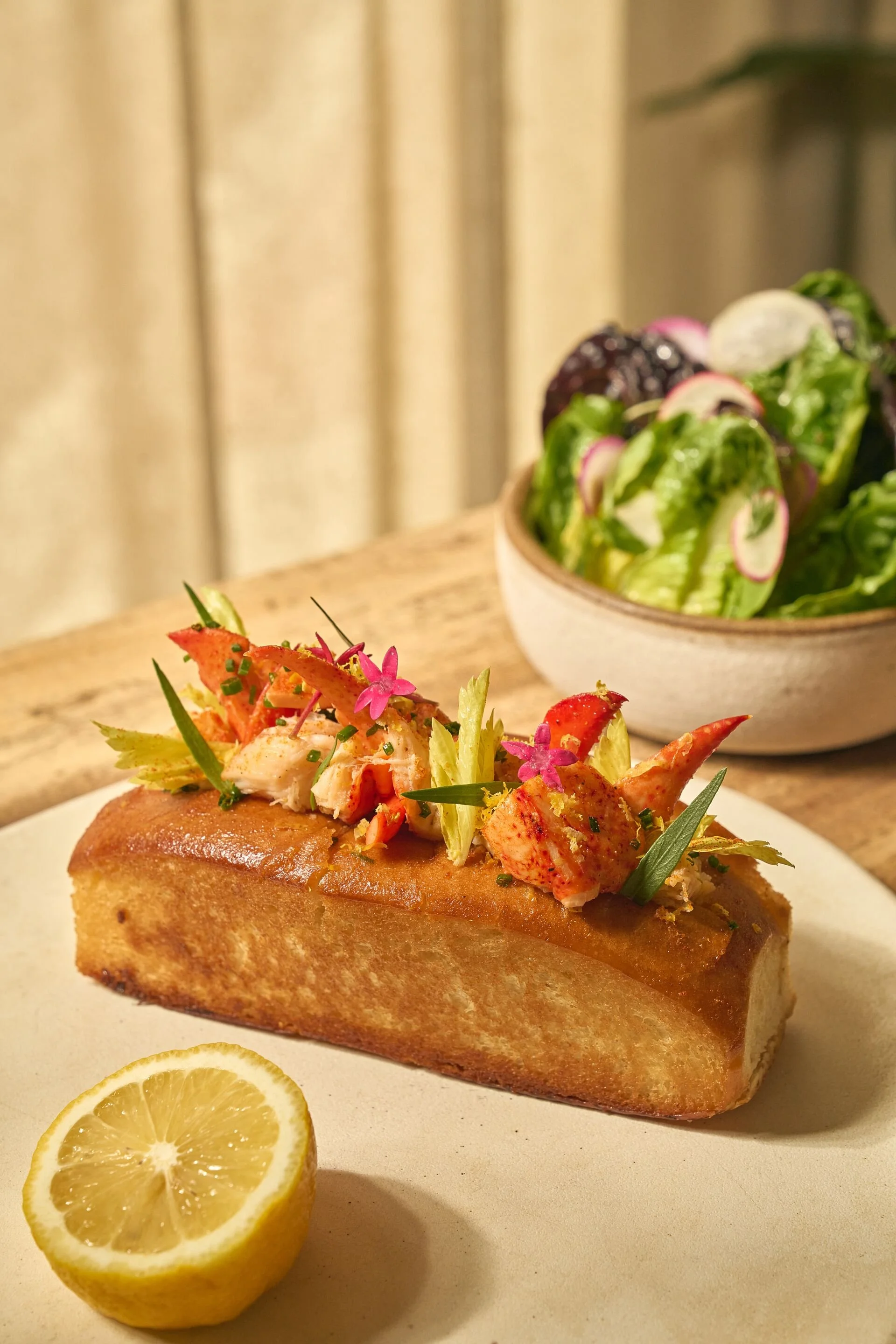 A slice of seafood loaf topped with lobster meat, garnished with herbs and edible flowers, with lemon slices on the side and a salad in a bowl in the background.