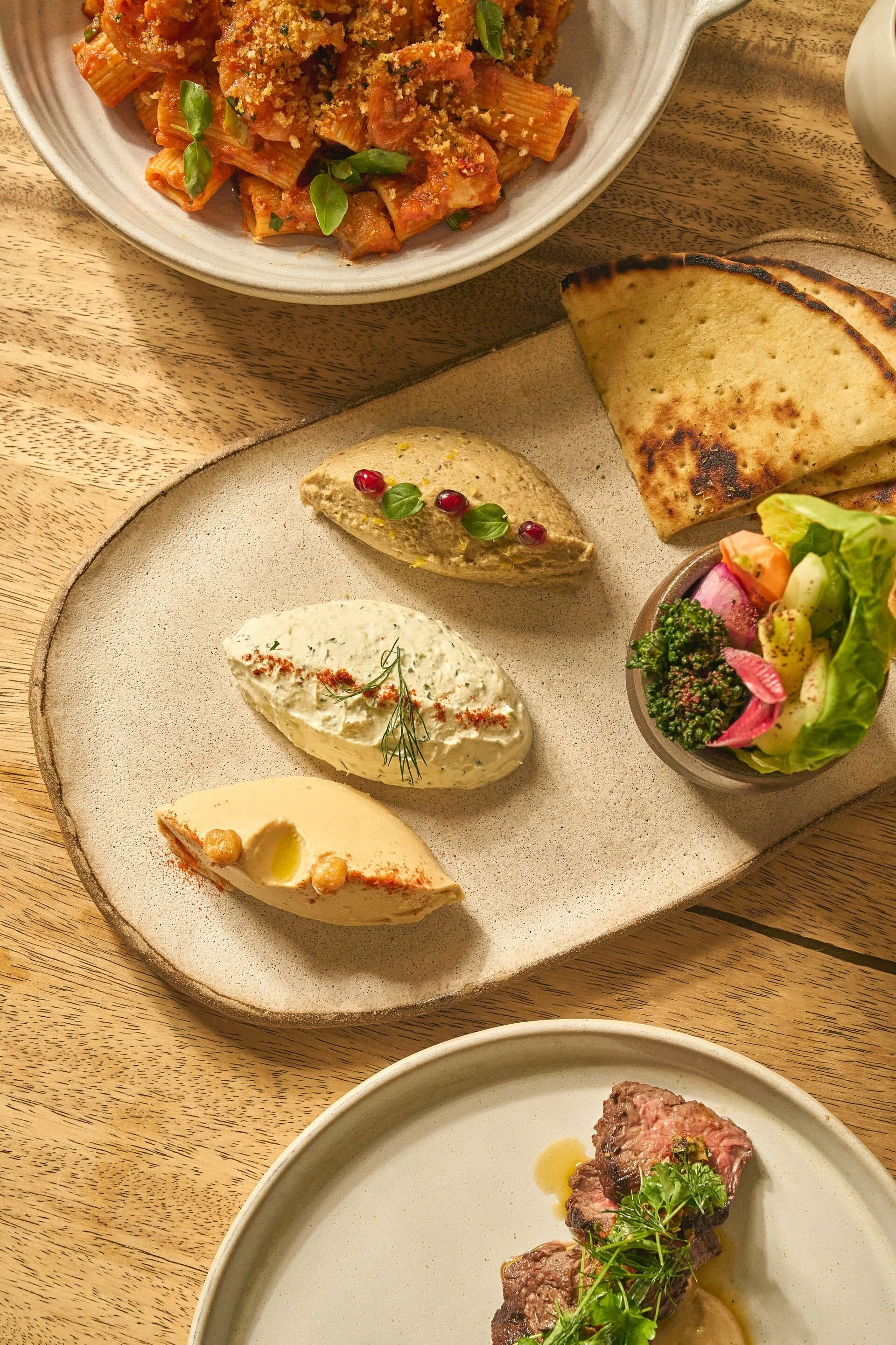 A plate of assorted appetizers including hummus with paprika and herbs, a slice of flatbread, a small salad with greens and vegetables, and some cheese with chickpeas and herbs, on a wooden table.