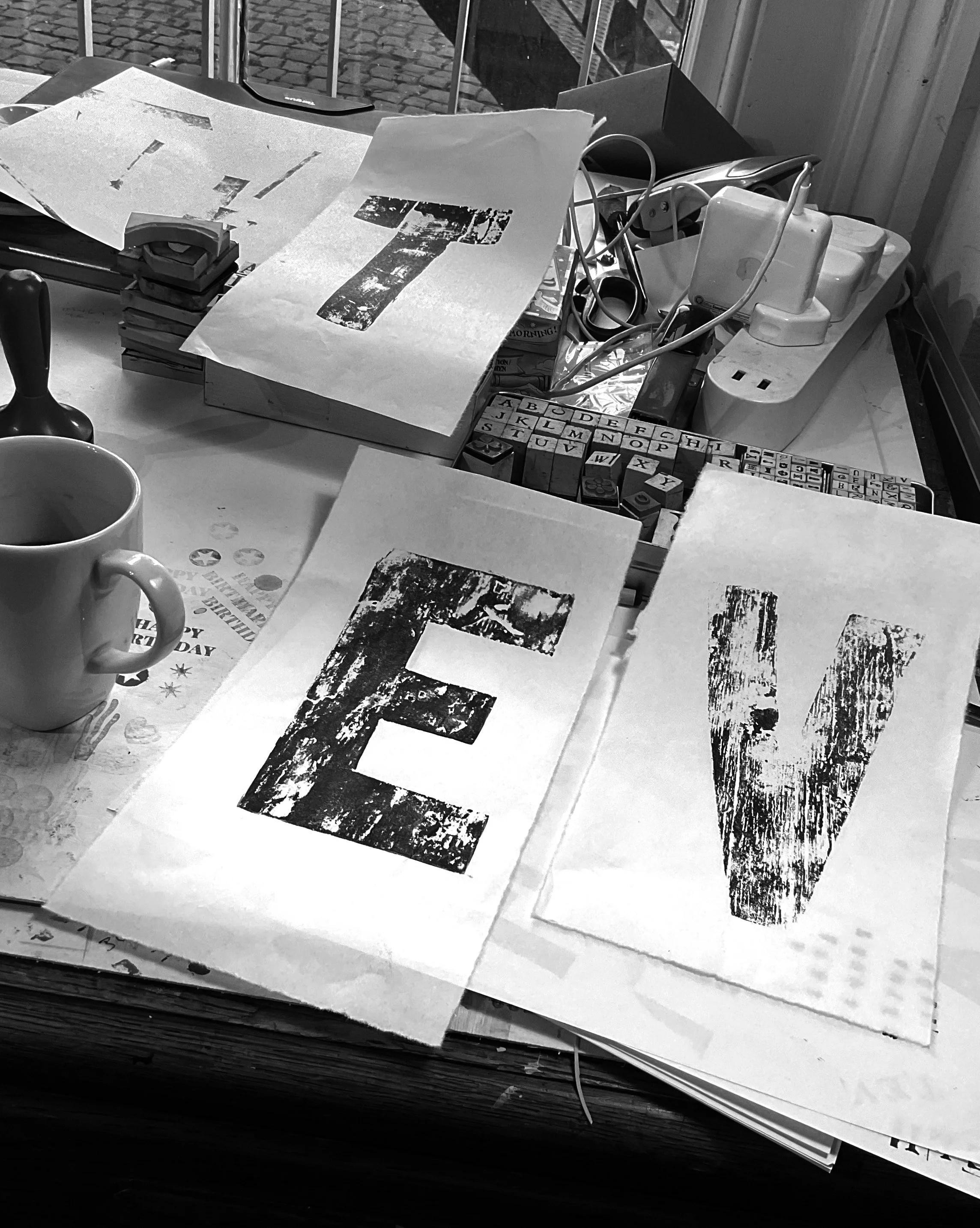 A cluttered desk with papers, a mug, rubber stamps, a power strip, cables, and various office supplies. The papers have large printed or stamped letters on them, and the background shows a window with a cobblestone street outside.