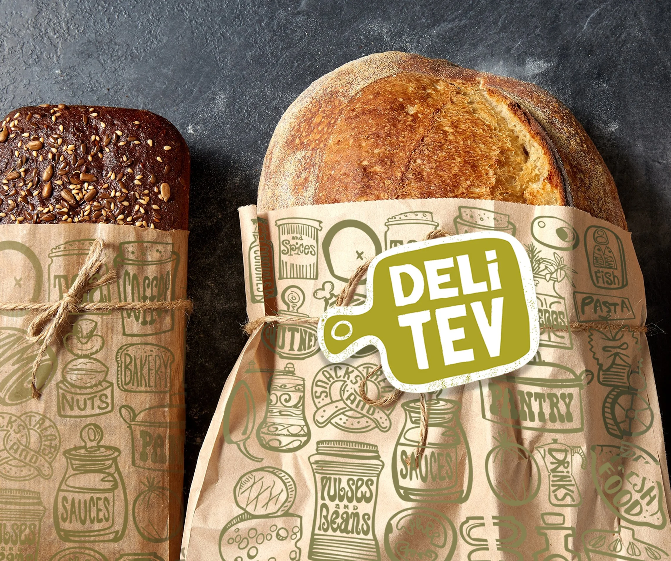 Close-up of two loaves of bread wrapped in paper with green illustrations and text, on a dark surface. One loaf is topped with seeds, the other has a golden crust and a partially sliced surface showing the interior.