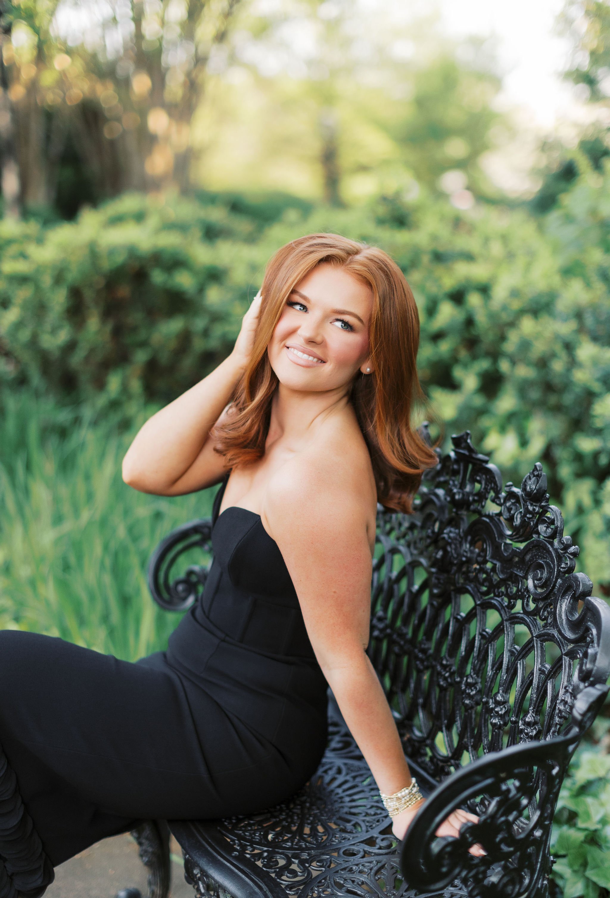 A woman with red hair and blue eyes sitting on a black ornate park bench in a green outdoor setting, smiling and touching her hair.