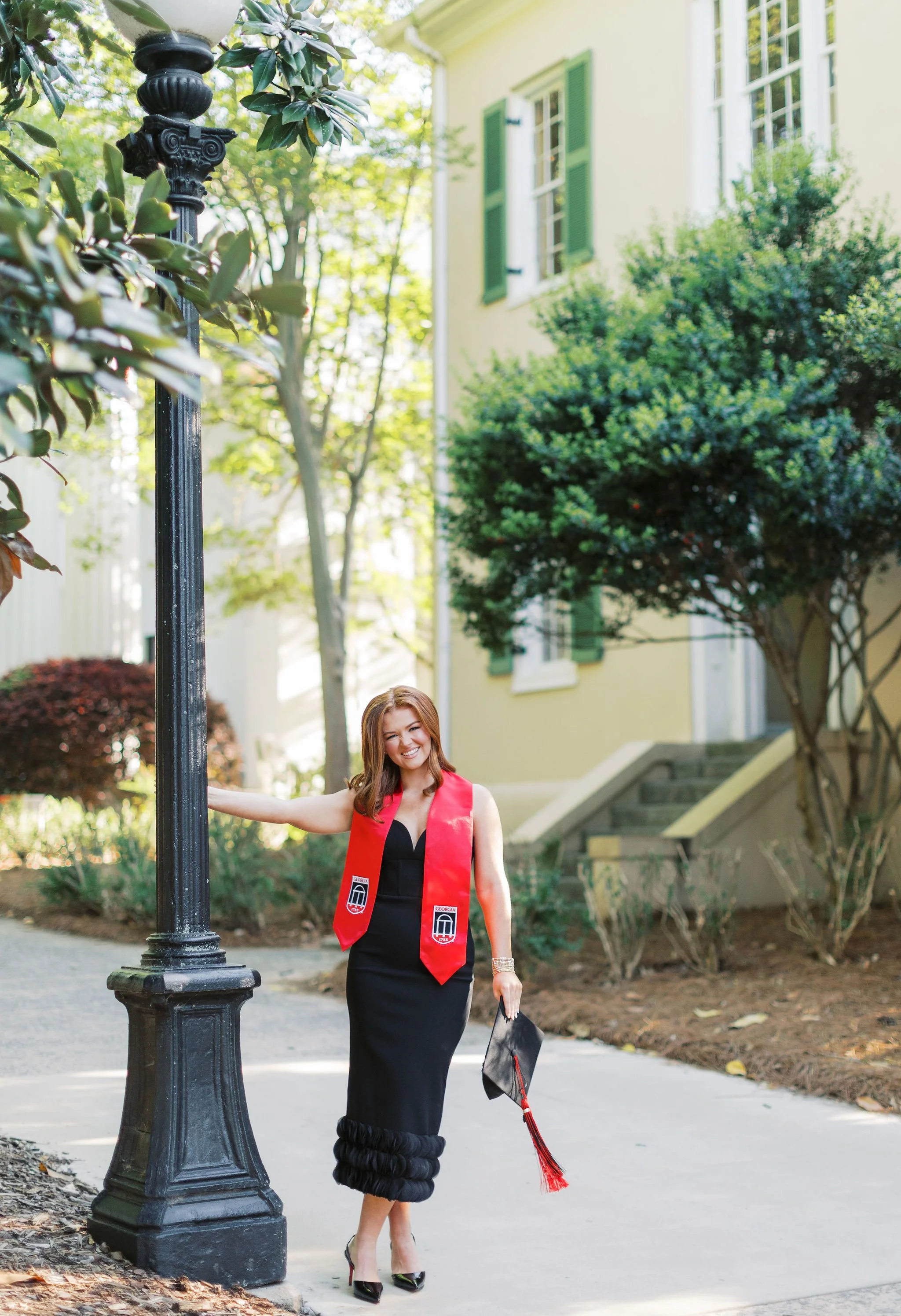 Woman in black dress and red graduation stole celebrating after graduation outside near a lamppost, trees, and a yellow house with green shutters.