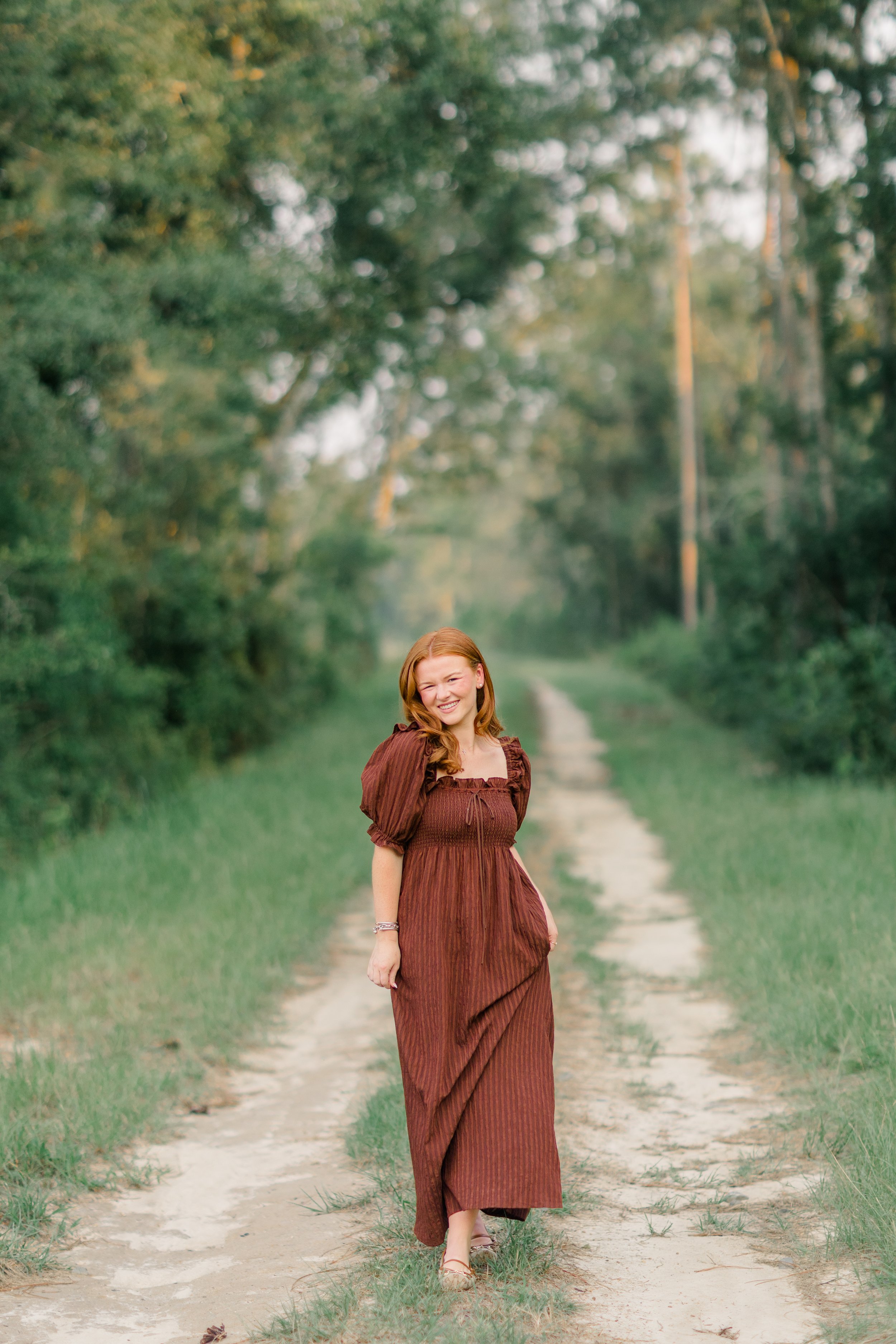 A young woman with red hair wearing a long, brown dress walking on a dirt path through a green, wooded area, smiling at the camera.