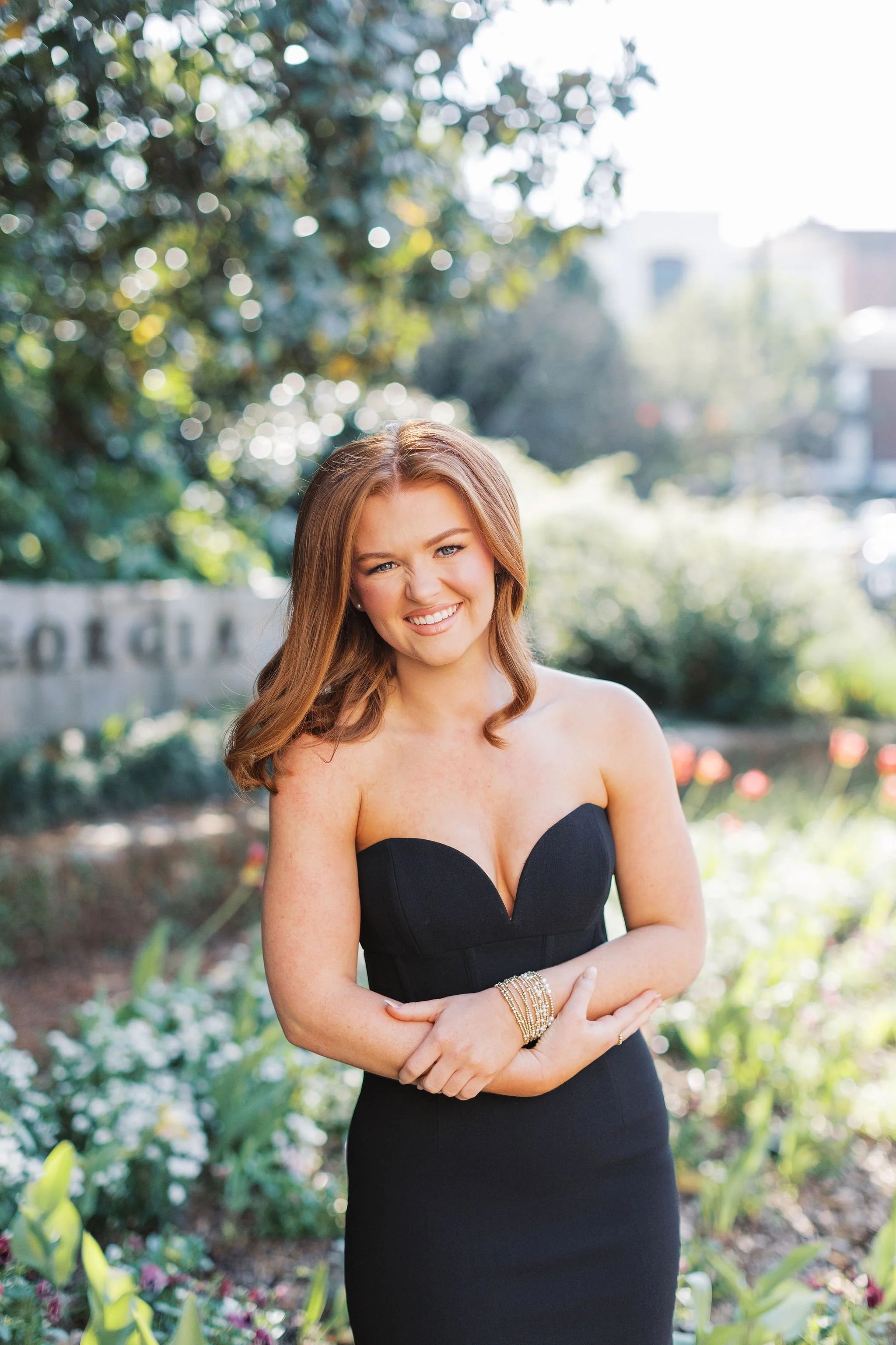 A woman with long, wavy brown hair wearing a strapless black dress standing outdoors in a garden with trees and flowers, smiling and crossing her arms.