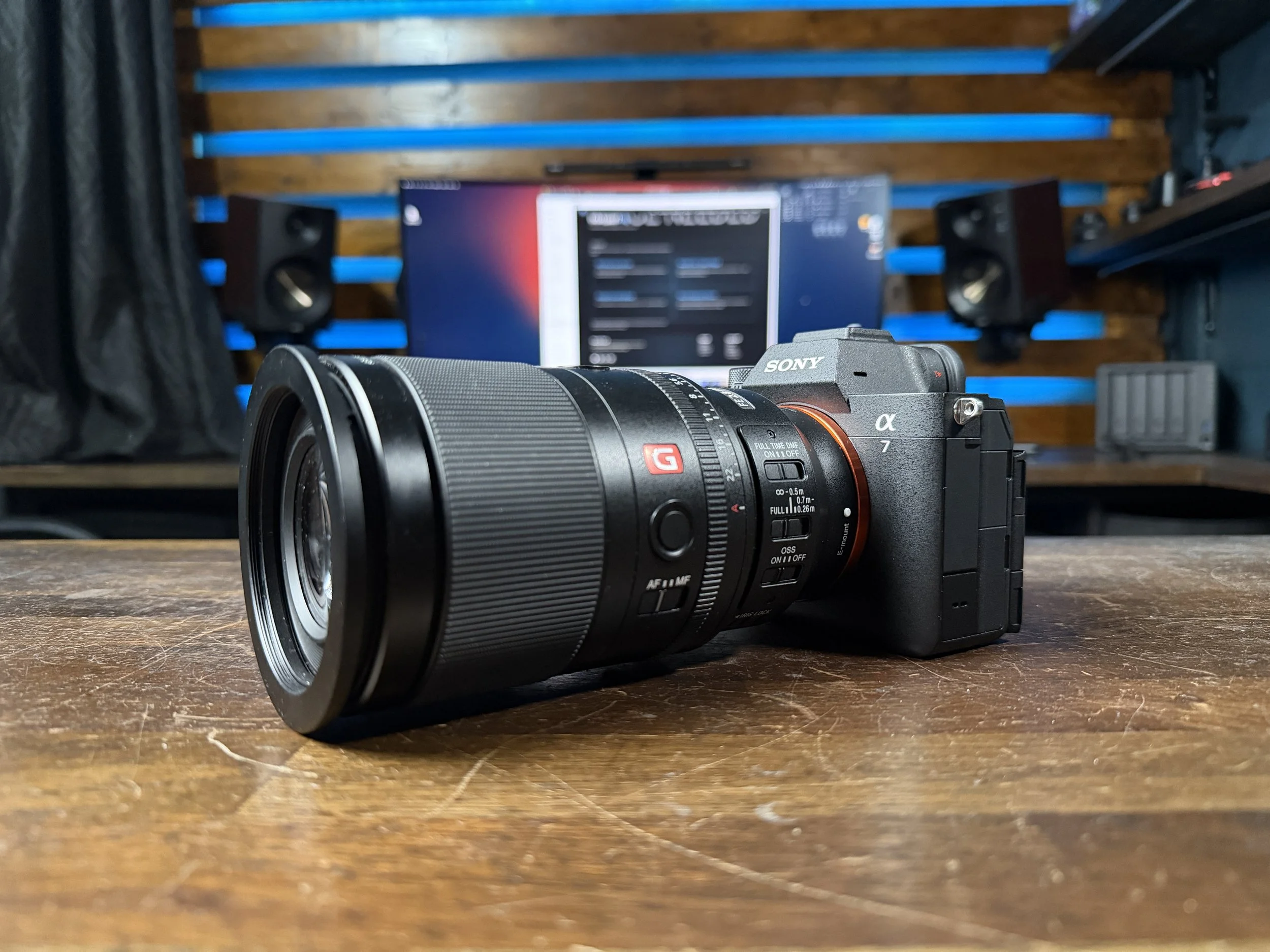 A Sony Alpha 7 camera with a zoom lens is placed on a wooden surface, with a blurred background featuring a computer monitor and speaker system mounted on a wood-paneled wall.