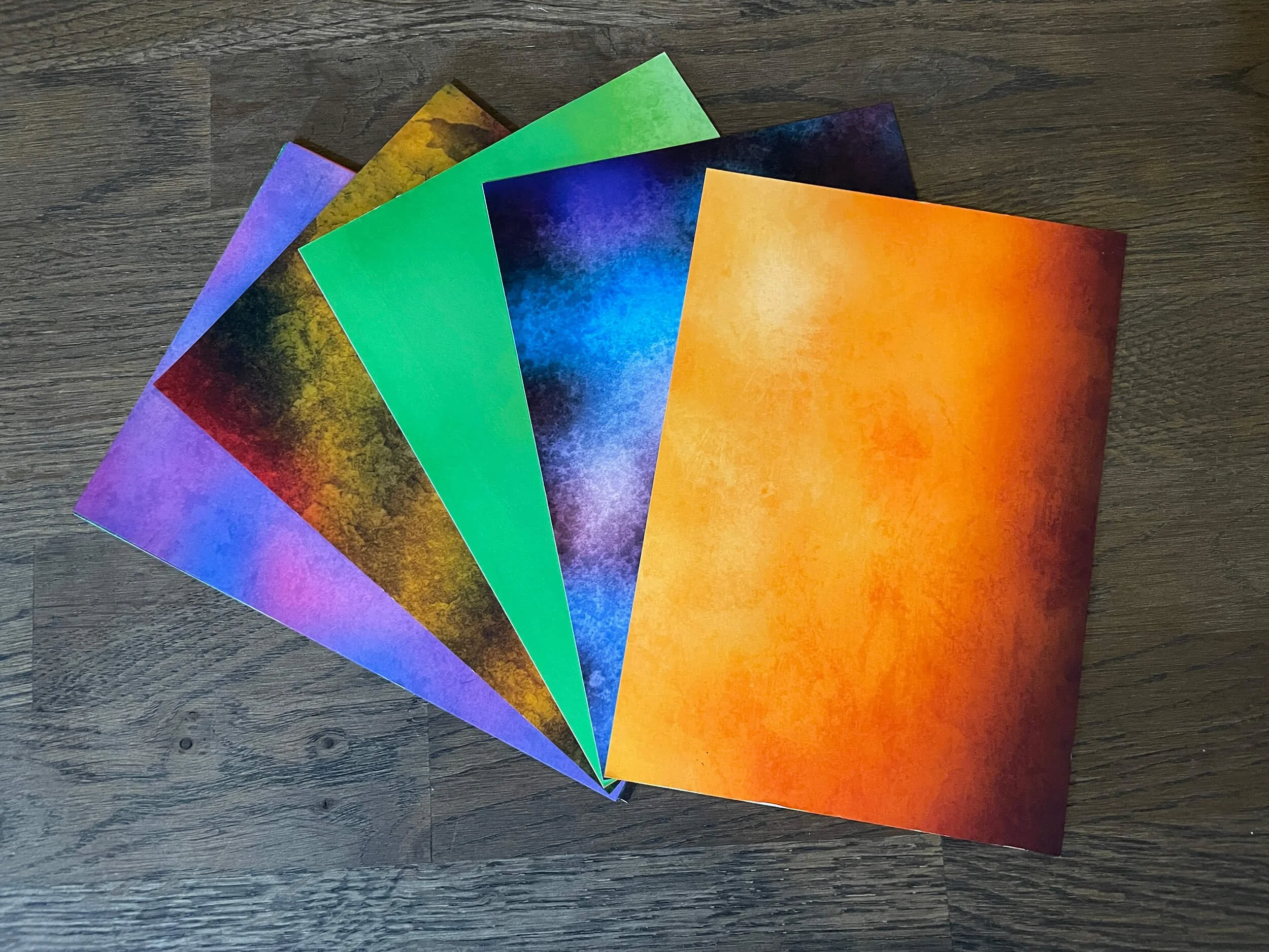 Colorful sheets of paper with galaxy-like patterns in purple, blue, green, yellow, orange, and red, fanned out on a dark wood surface.