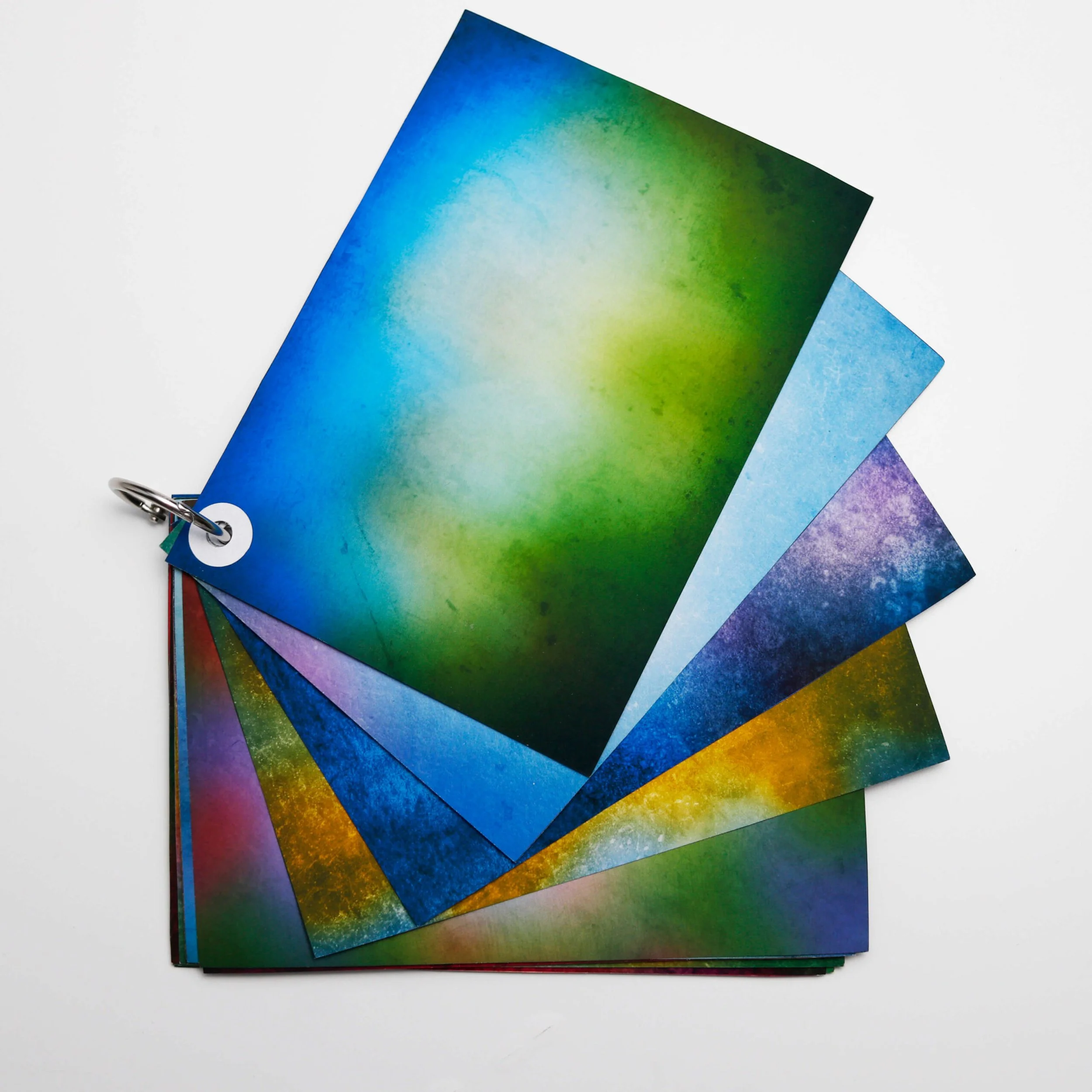 Stack of colorful watercolor postcards or prints in shades of blue, green, purple, and yellow, fanned out on a white surface.