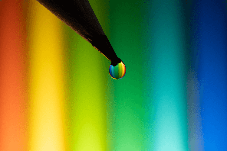 Close-up of a rainbow-colored water droplet hanging from the tip of a black tool, with a vibrant rainbow background.