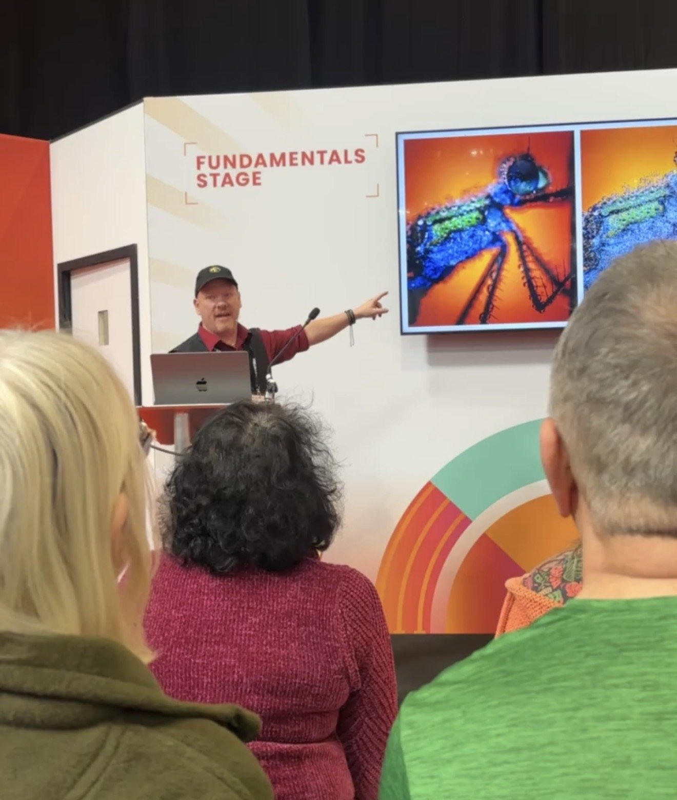 A man wearing a black cap and red shirt giving a presentation at the Fundamentals stage, pointing to a colorful poster of a dragonfly on a screen, with an audience of diverse people watching.