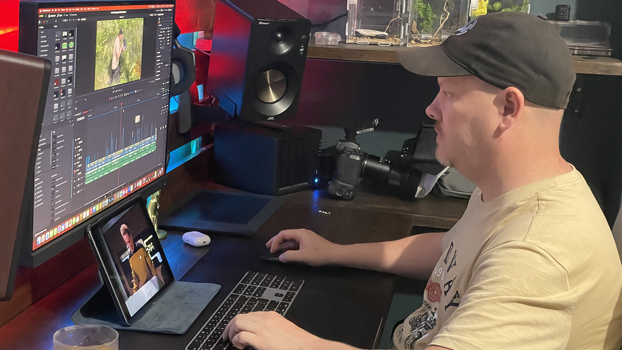 A man wearing a black cap and beige T-shirt working at a computer desk with a large monitor displaying video editing software, a camera, and an iPad showing a person on stage.