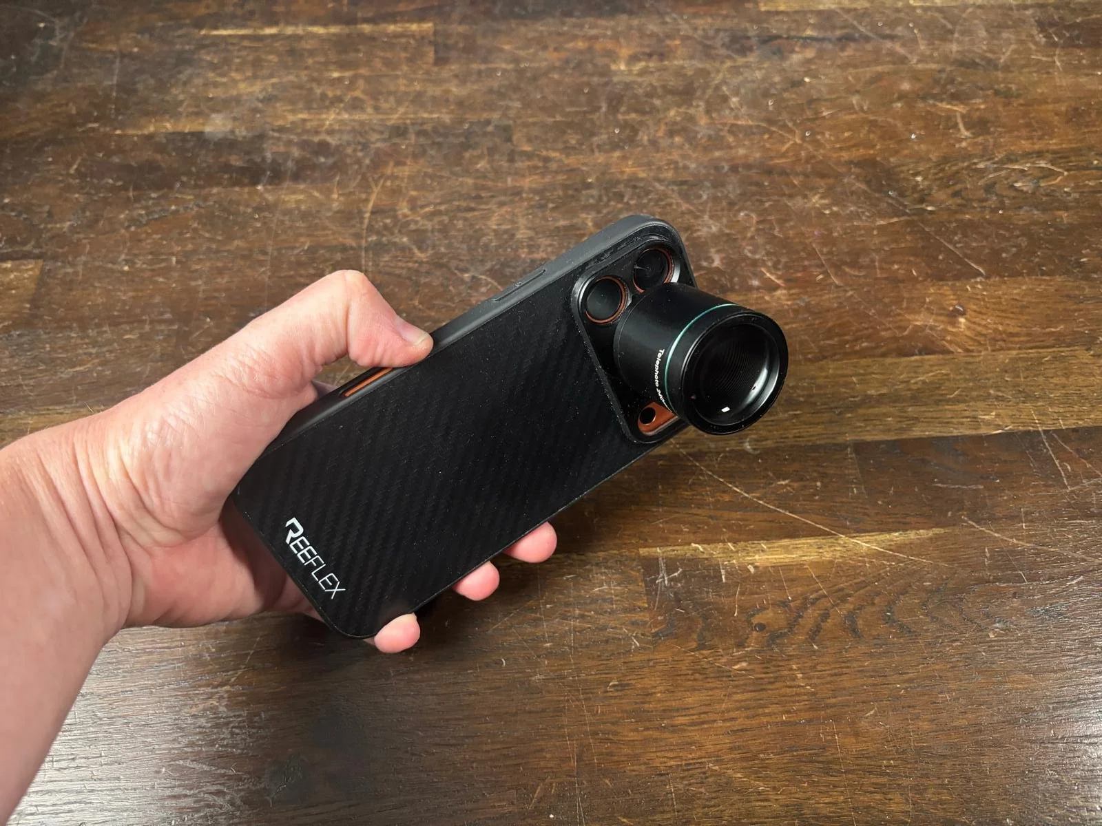 A hand holding a REELIX camera with a large lens and multiple small lenses on the front, against a wooden surface.