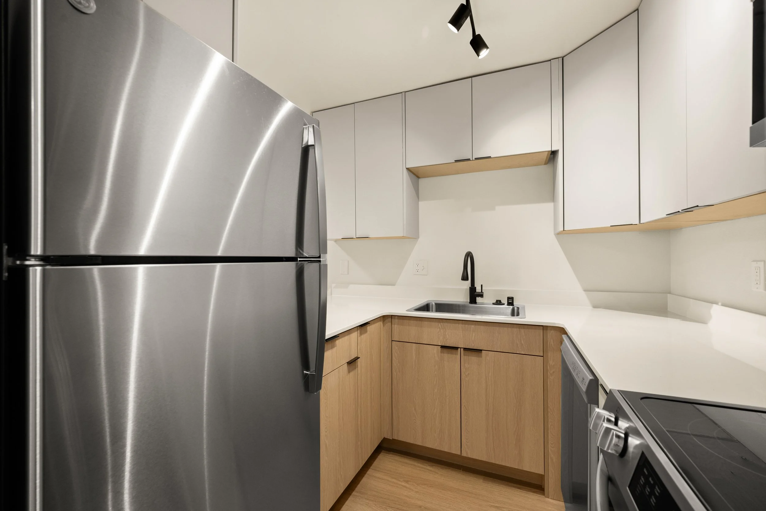 Modern kitchen with stainless steel refrigerator, white countertops, light wood cabinets, black sink faucet, and a washing machine, under black ceiling light fixtures.
