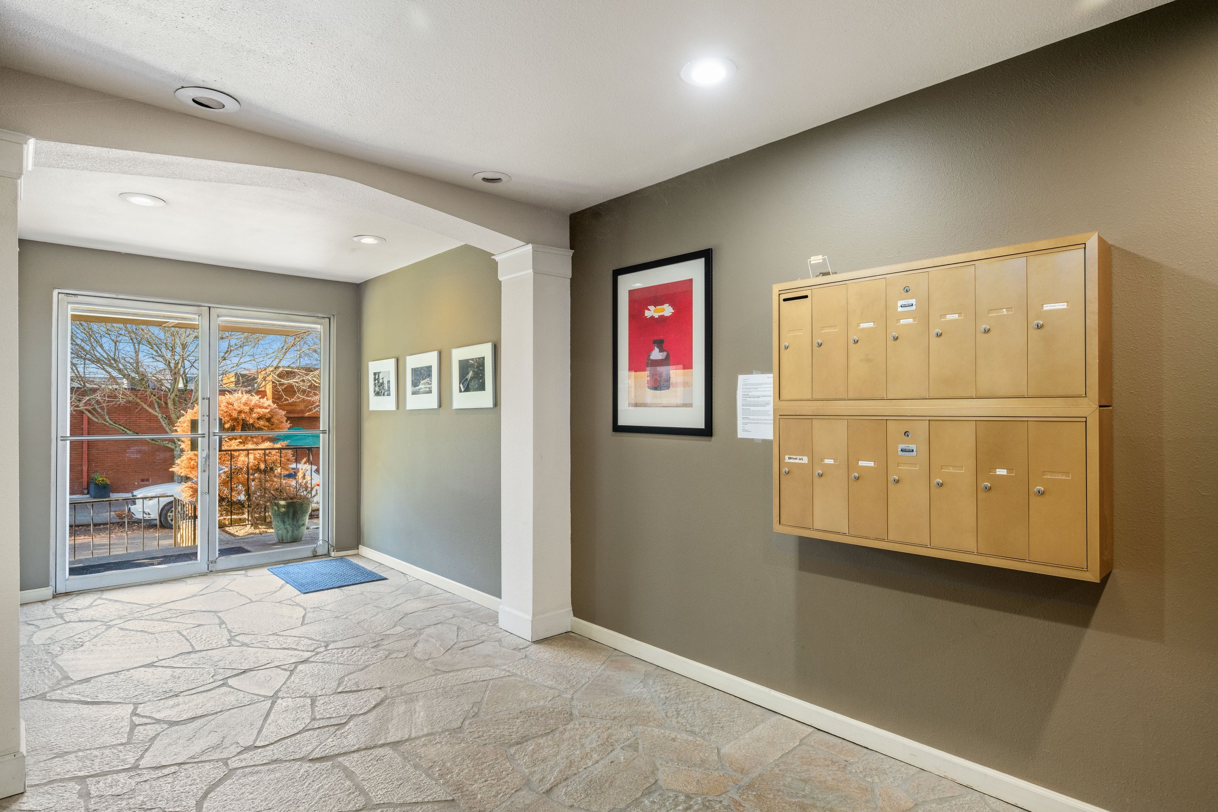 Indoor hallway with a glass door leading outside, wooden mailboxes on the right wall, framed artwork and photo prints on the wall, and a stone floor.