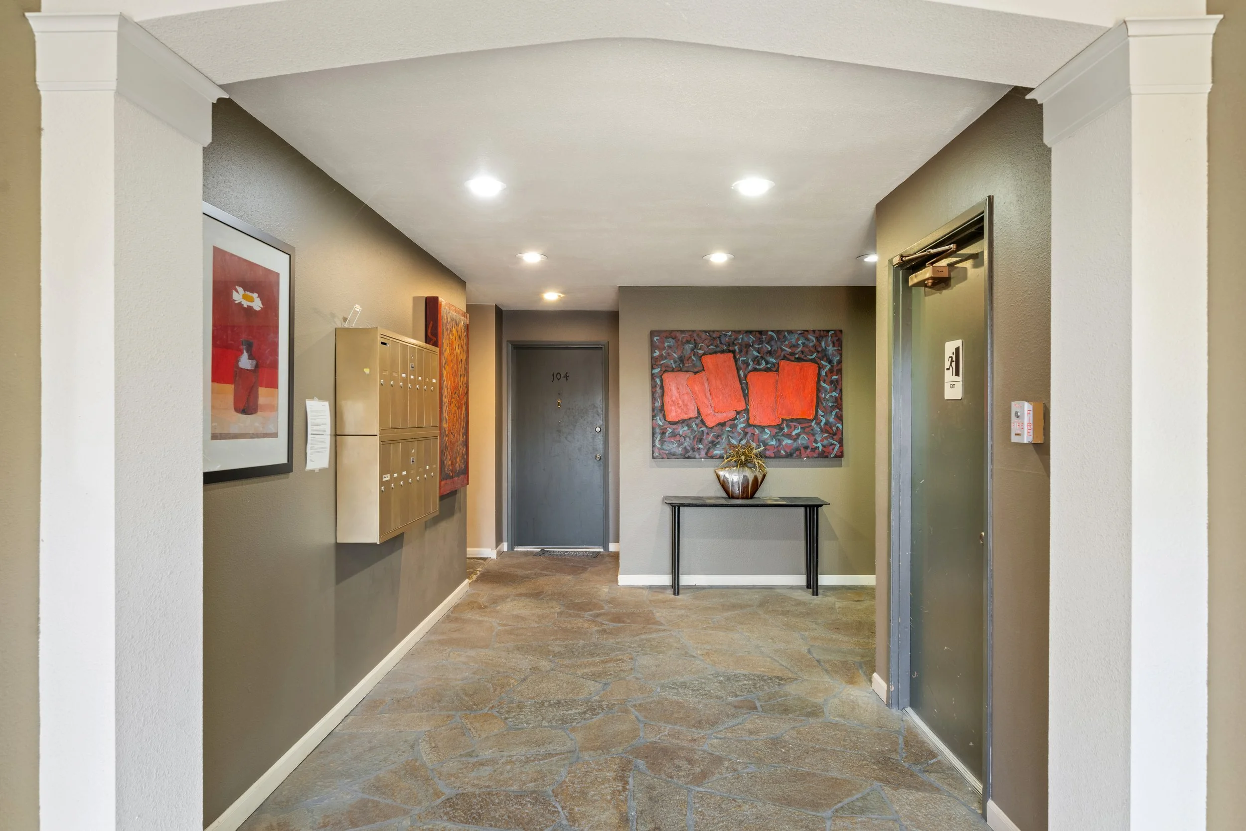 Indoor apartment building lobby with a gray door numbered 104, a desk with a decorative vase on top, artwork on the walls, and a staircase with a sign indicating an elevator.