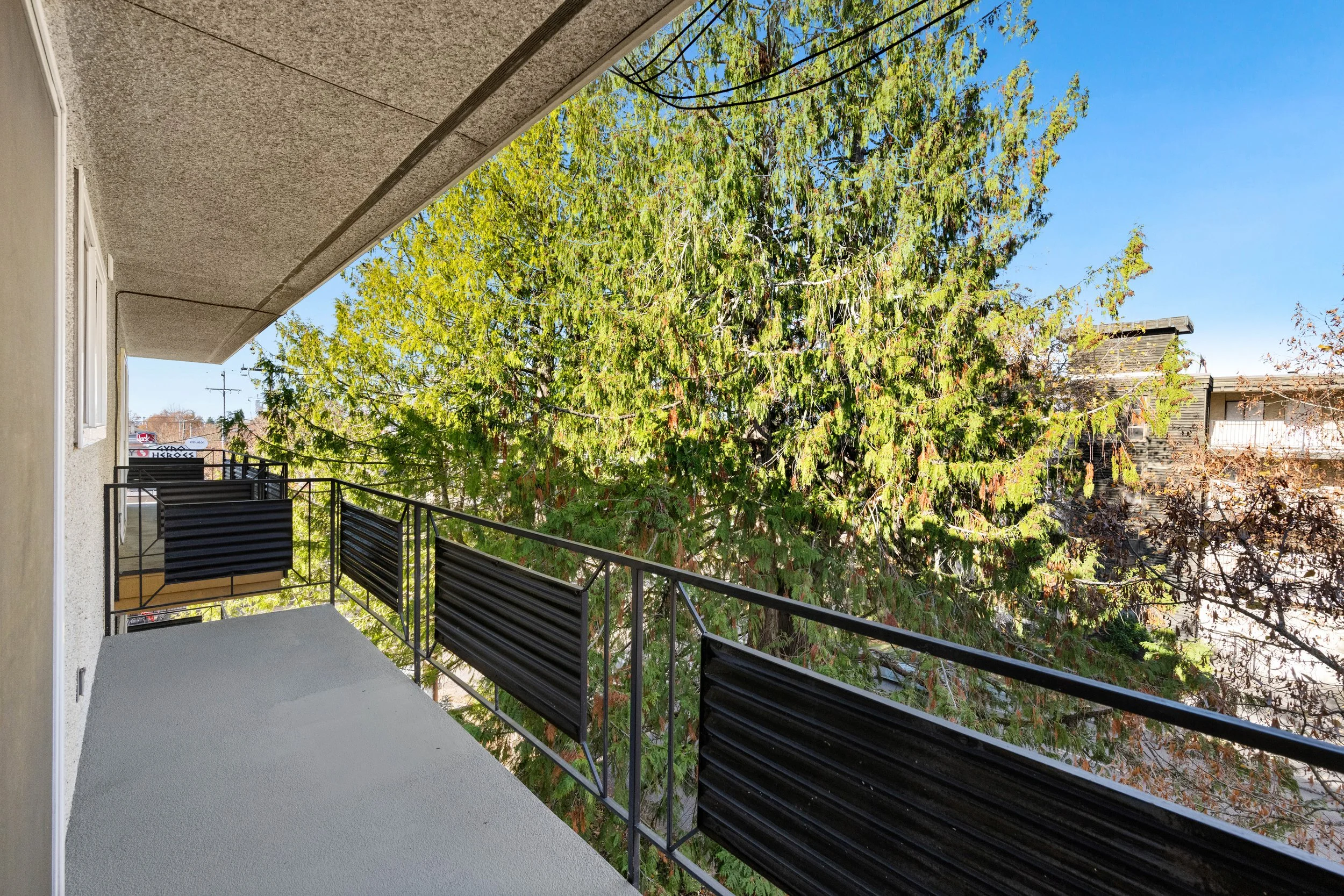 Balcony with metal railings and black planters, overlooking tall green trees and neighboring buildings, under a partly cloudy blue sky.
