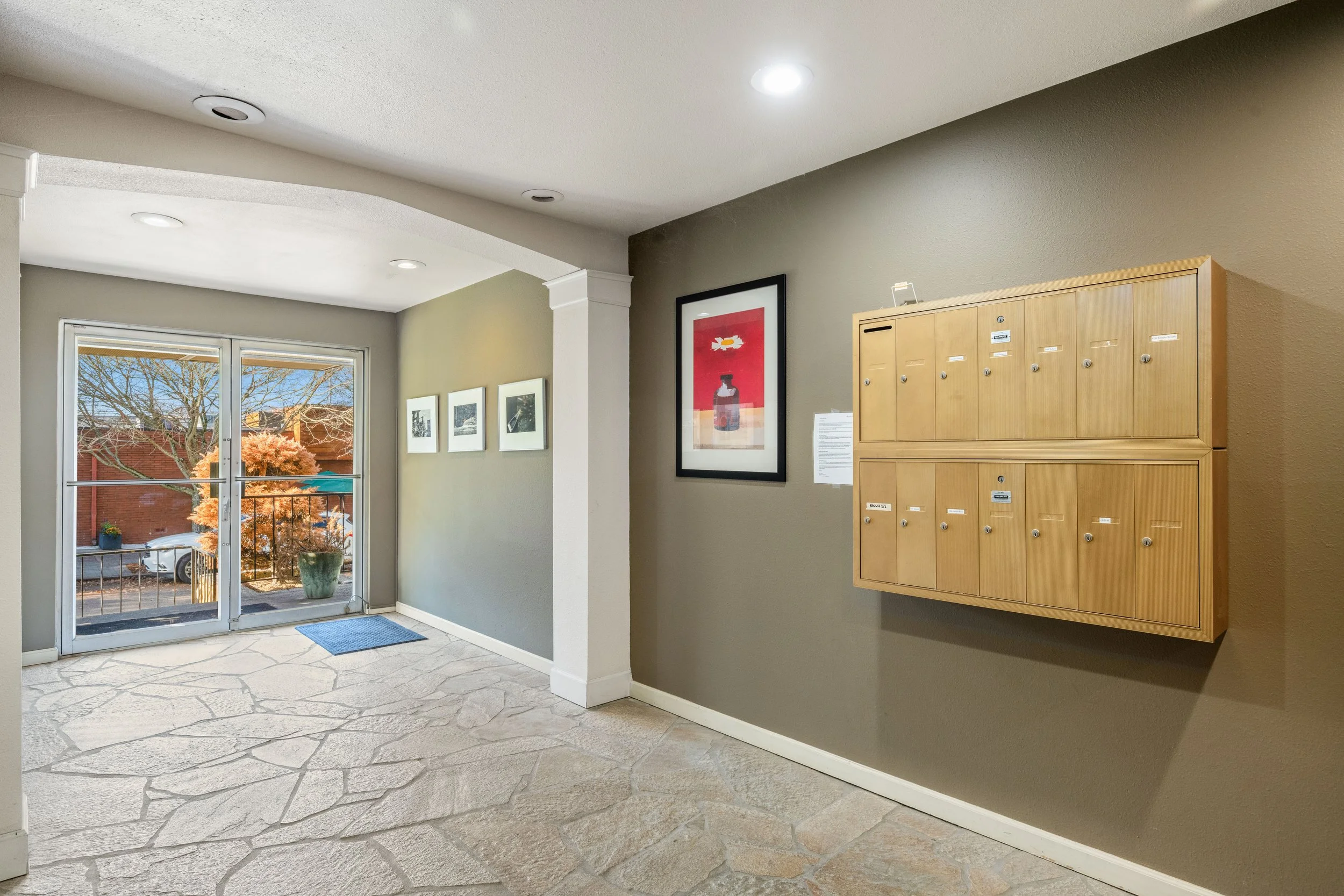 Apartment lobby with glass sliding door, framed artwork on walls, wood mailboxes, and stone floor.