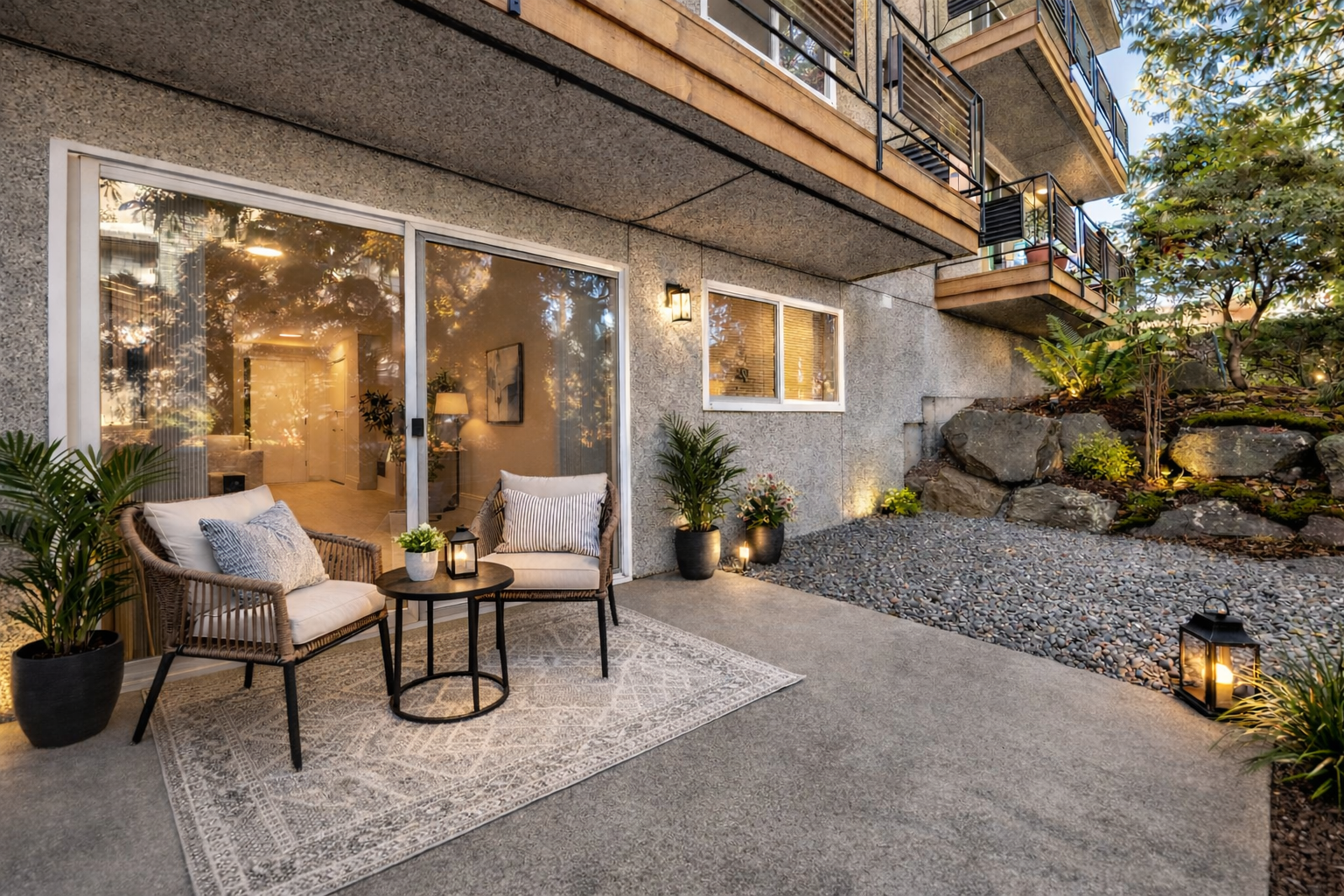 An outdoor patio area with two wicker chairs, a round table, and potted plants, decorated with lanterns at dusk, with a sliding glass door leading inside a house.