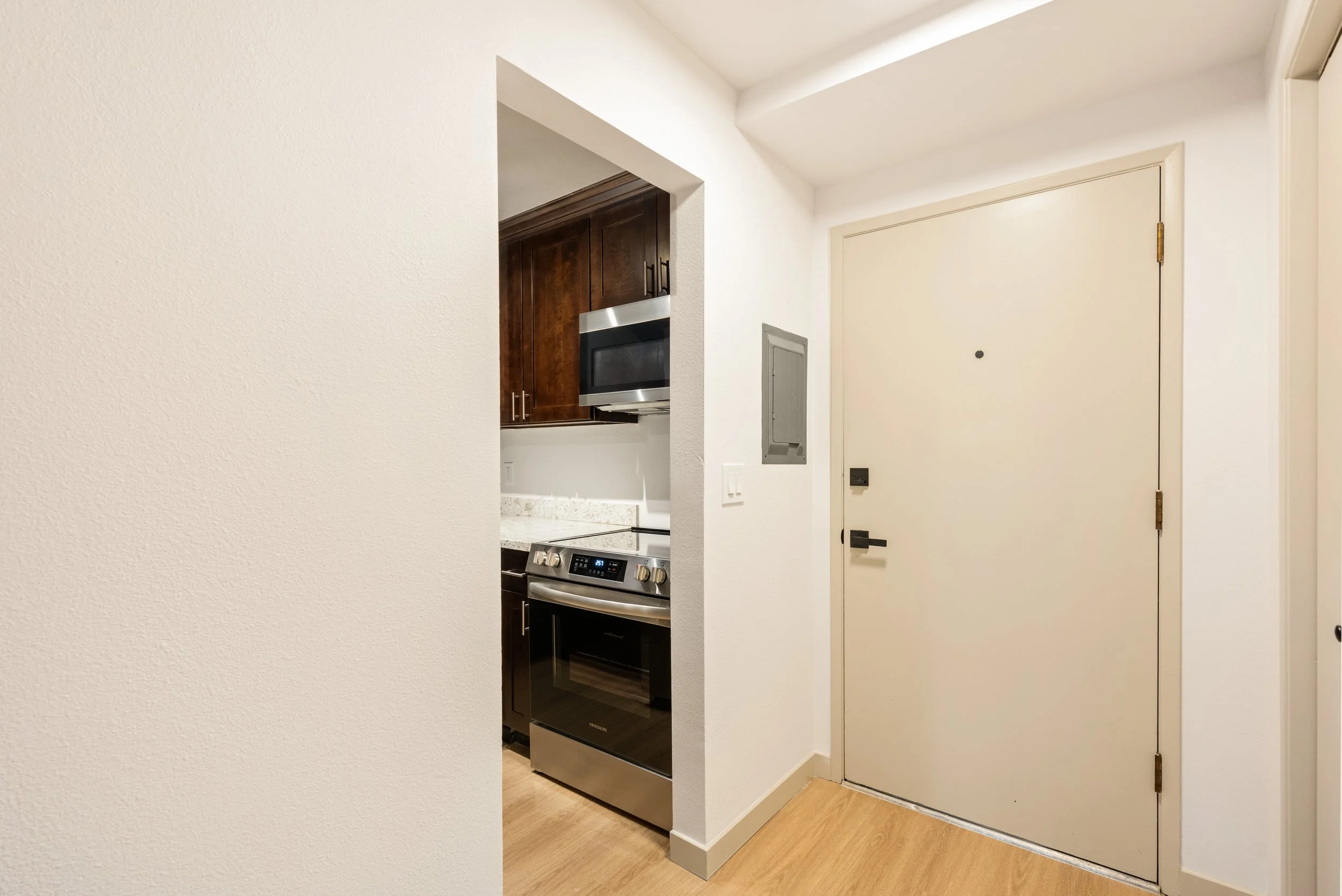 Interior view of an apartment foyer showing a white door, a small electrical panel on the wall, and part of a kitchen with dark wooden cabinets, a microwave, and a stainless steel stove.