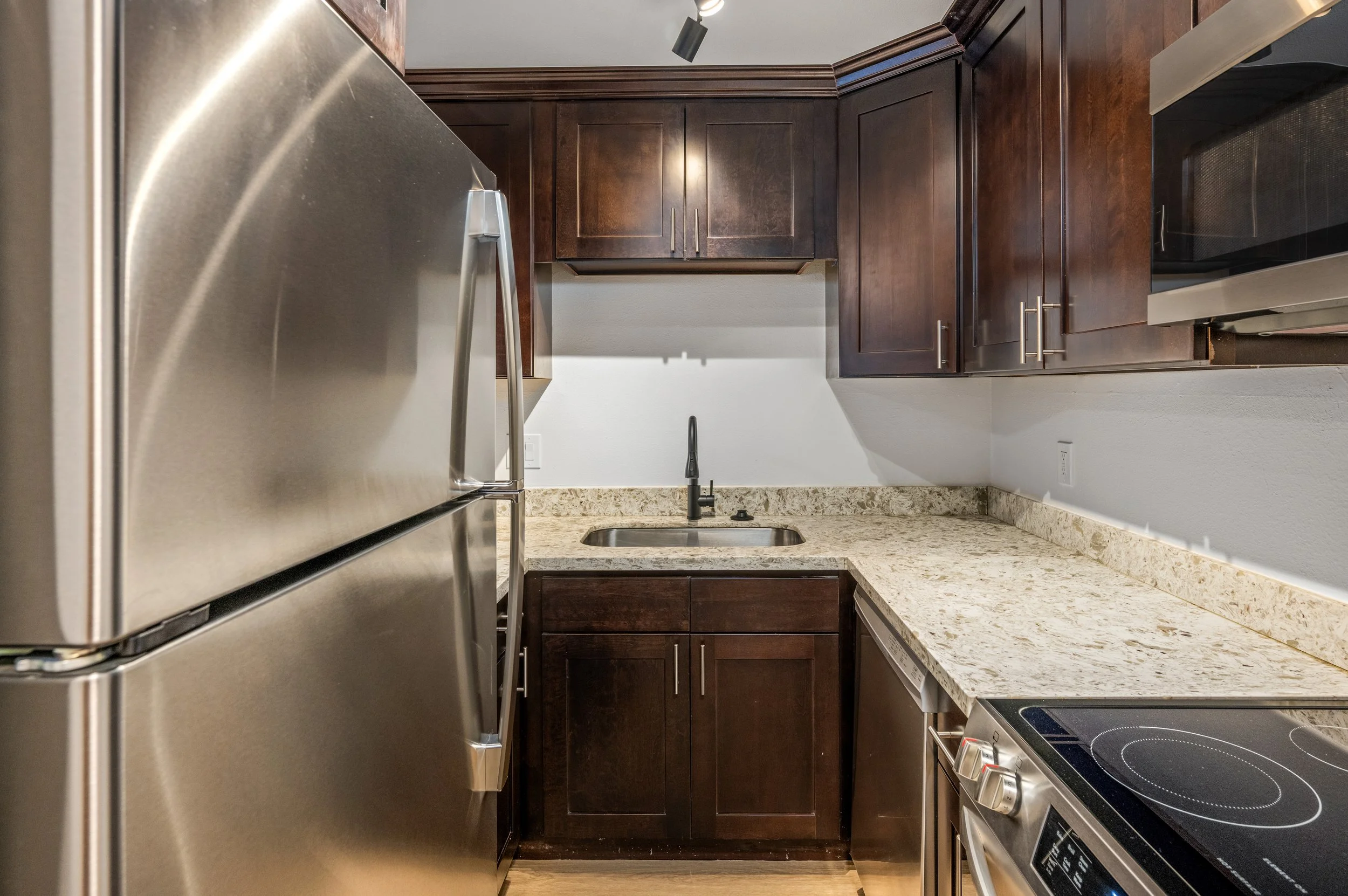 Kitchen with dark brown wooden cabinets, a granite countertop, a stainless steel refrigerator on the left, a small sink in the center, and a stainless steel oven on the right.